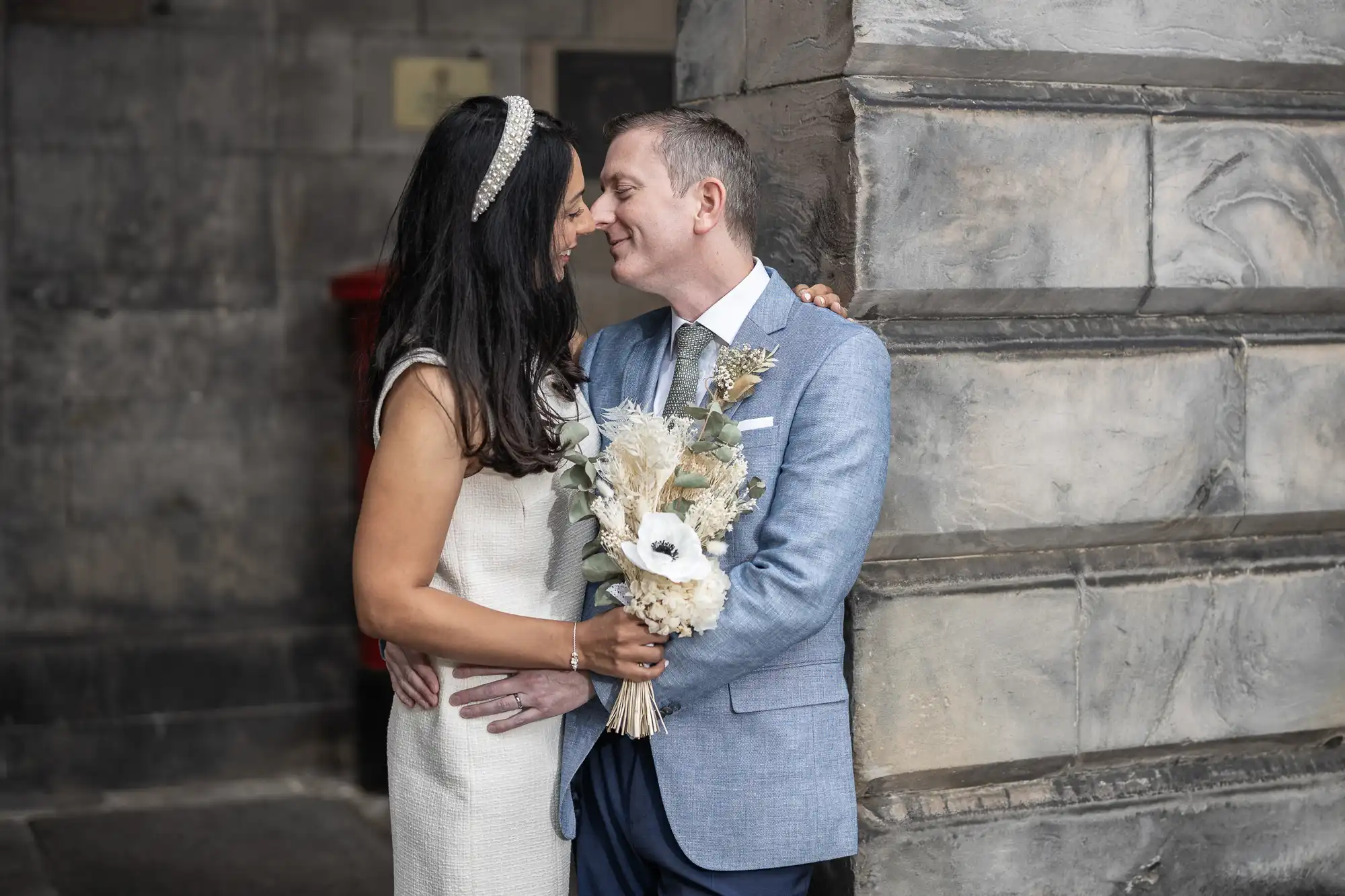 A couple in wedding attire embraces by a stone wall, the woman holding a bouquet of white and beige flowers.