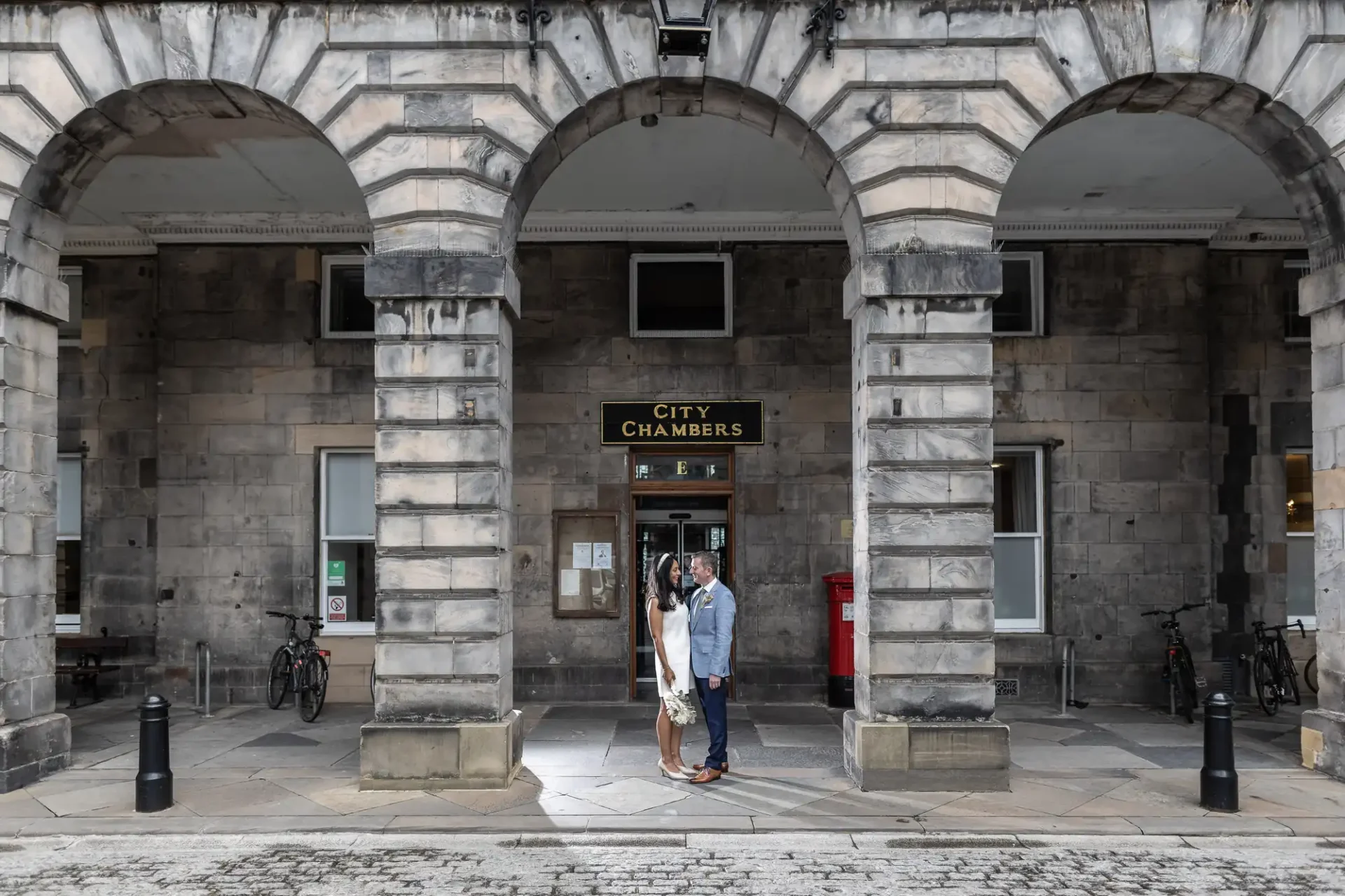 A couple stands facing each other under the arches in front of the entrance to a stone building labeled "City Chambers." The area is paved with cobblestones, and bicycles are parked nearby.