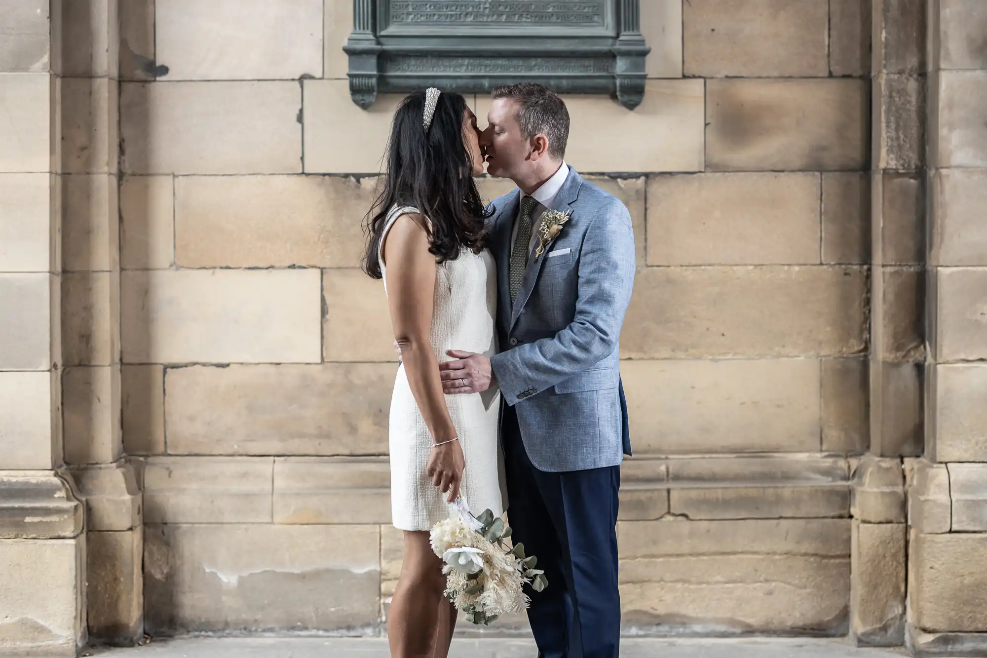 A couple dressed in wedding attire kisses in front of a stone wall. The woman holds a bouquet of flowers.
