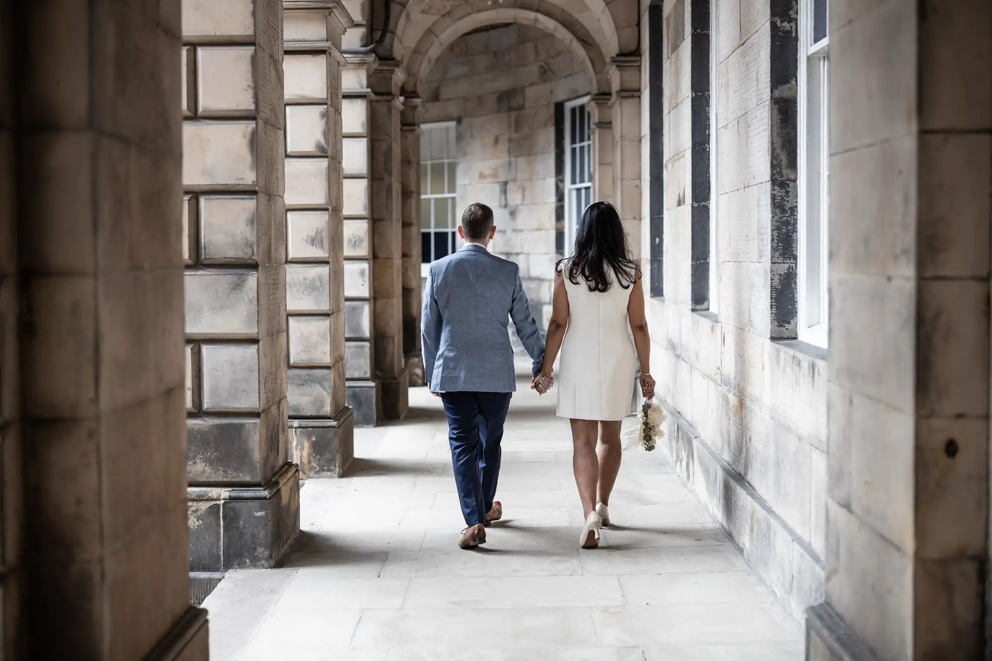 A couple holding hands walks down a stone corridor with high arched ceilings. The man is wearing a blue suit and the woman is in a white dress, holding a small bouquet.