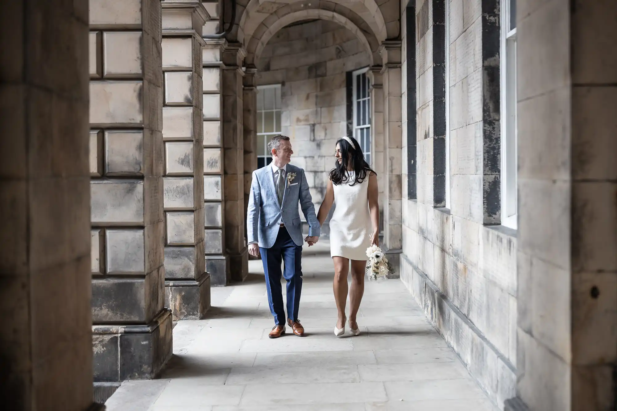 A couple is walking hand in hand through a stone corridor. The man is wearing a light blue suit, and the woman is wearing a short white dress, holding a small bouquet of flowers.
