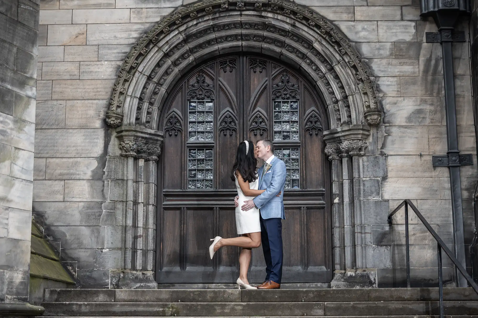 A couple shares a kiss in front of an ornate, arched wooden door of a stone building. The woman is lifting one leg and wearing a white dress, while the man is in a light blue blazer and dark pants.