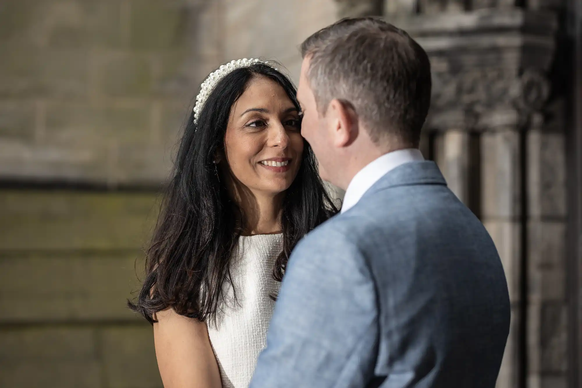 A woman in a white dress and a man in a light blue suit gaze at each other and smile in front of a stone building.