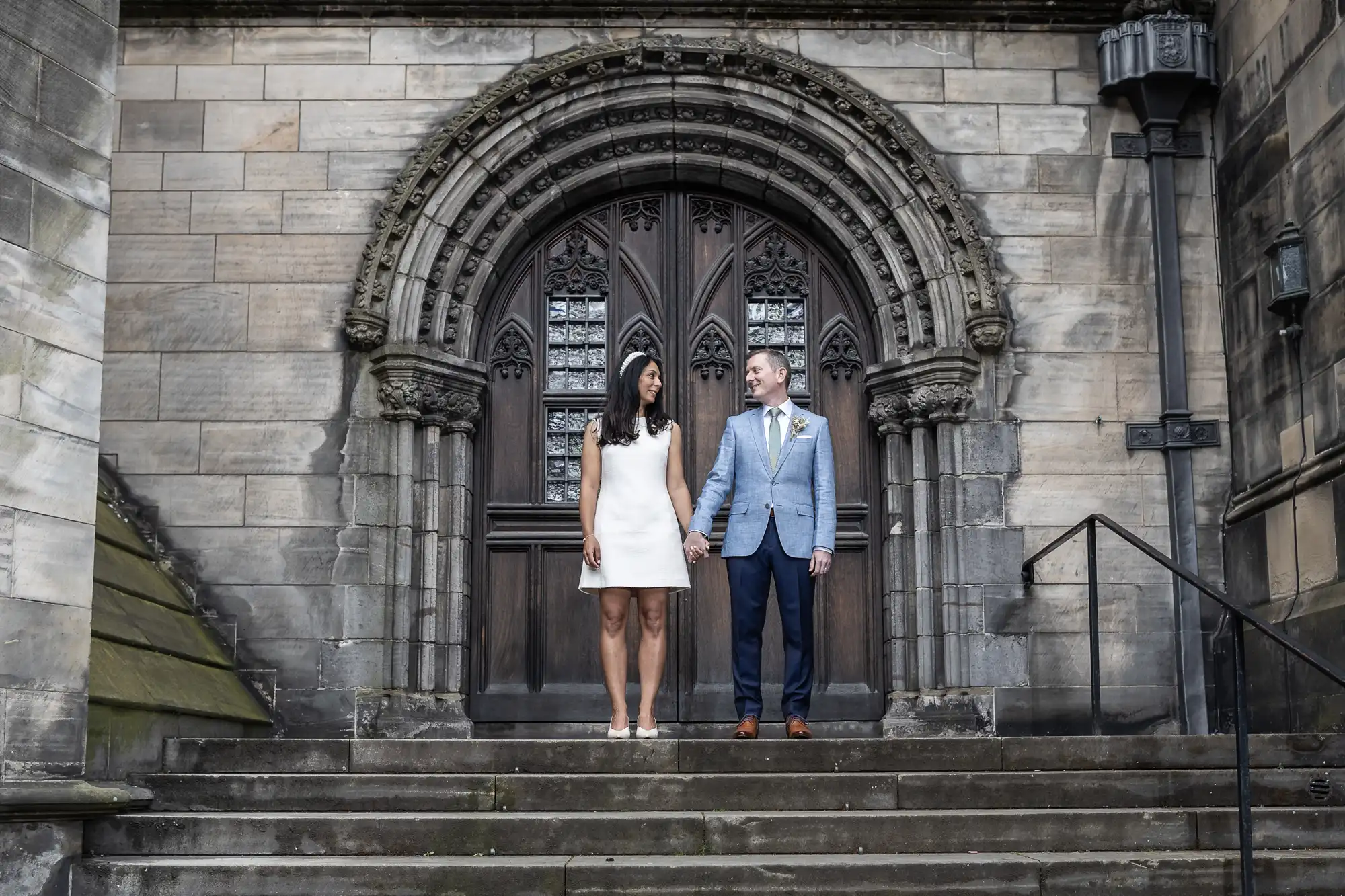 A couple stands hand in hand on stone steps in front of an ornate, arched wooden door set in a stone wall. The woman wears a white dress; the man wears a light blue jacket and dark trousers.