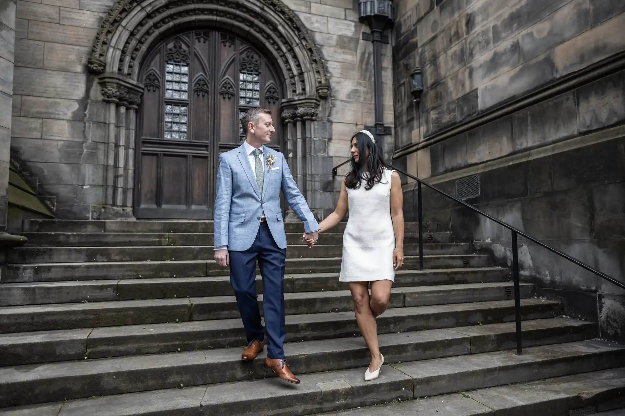 A couple, dressed formally, walks hand in hand down stone stairs outside a historic building with arched wooden doors.