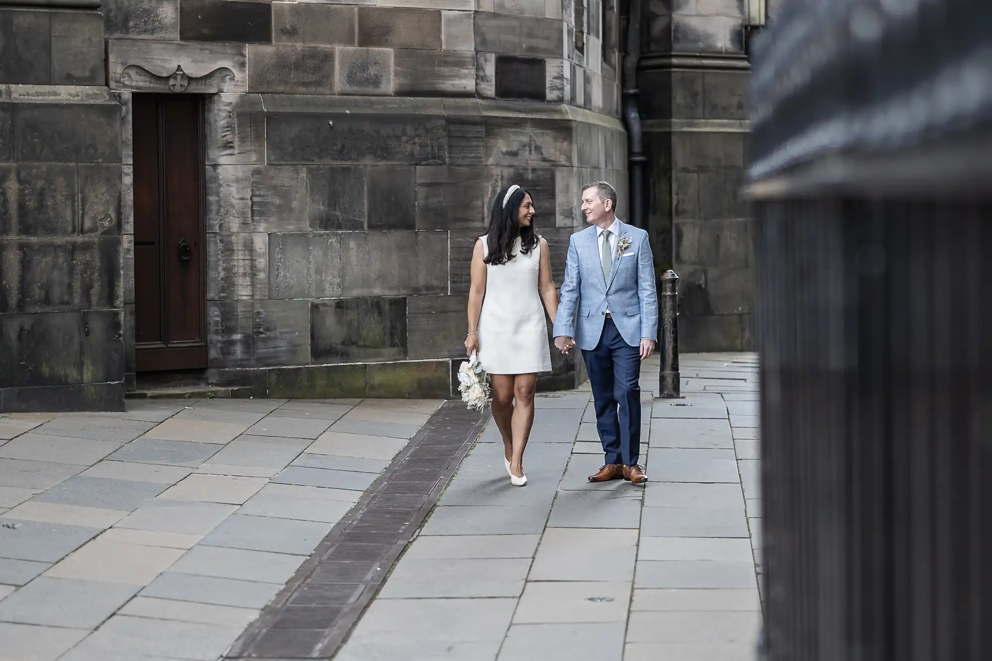 A couple holding hands walks on a stone pavement near a historic building. The woman wears a white dress and holds flowers, while the man is in a light blue suit.