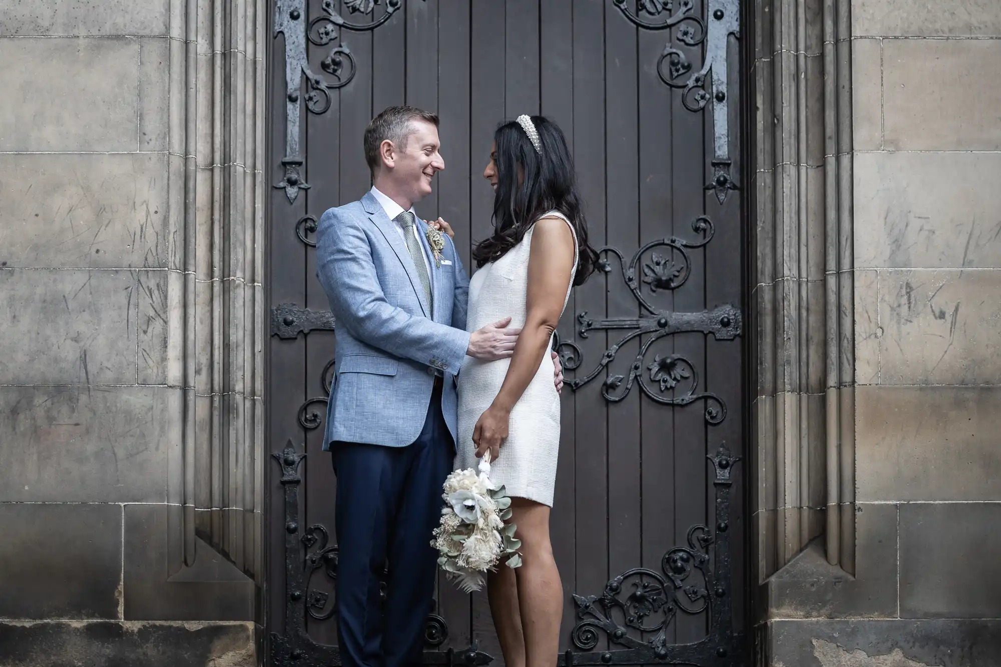 A couple stands facing each other in front of a large wooden door with intricate metalwork. Both are dressed in formal attire; the woman holds a bouquet of flowers.