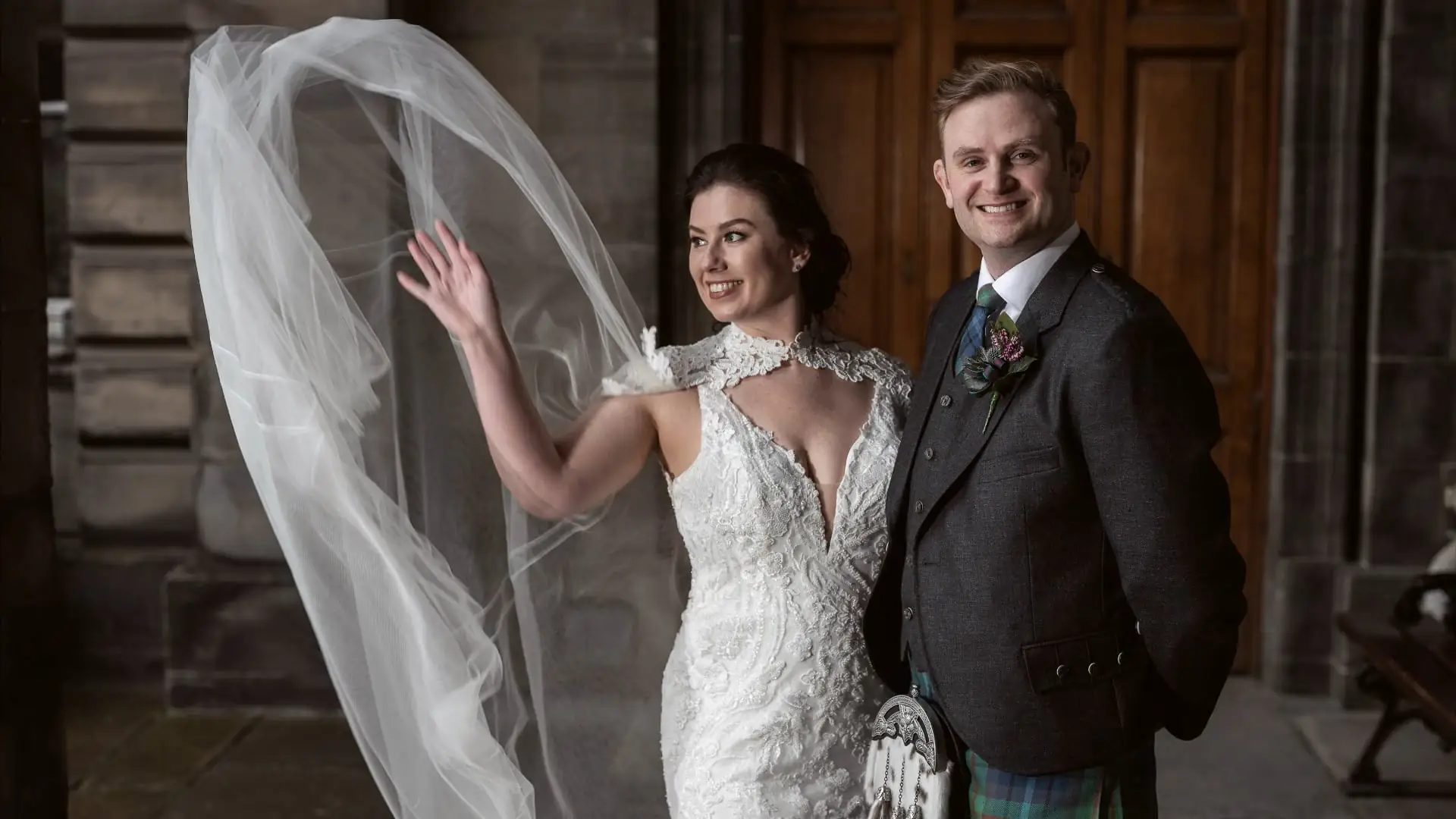 A bride in a white gown waves as her veil flows in the air, standing next to a smiling groom in a suit with a tartan kilt, in front of wooden doors.