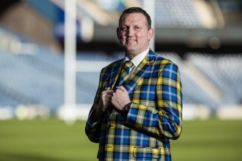 Product photography with Doddie Weir in a blue and yellow plaid suit stands in a sports stadium, smiling with his hands adjusting his jacket.