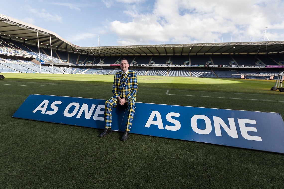 A person in a colorful plaid suit sits on a large sign that reads "AS ONE" in a stadium. The stadium is empty, and the sky is partly cloudy.