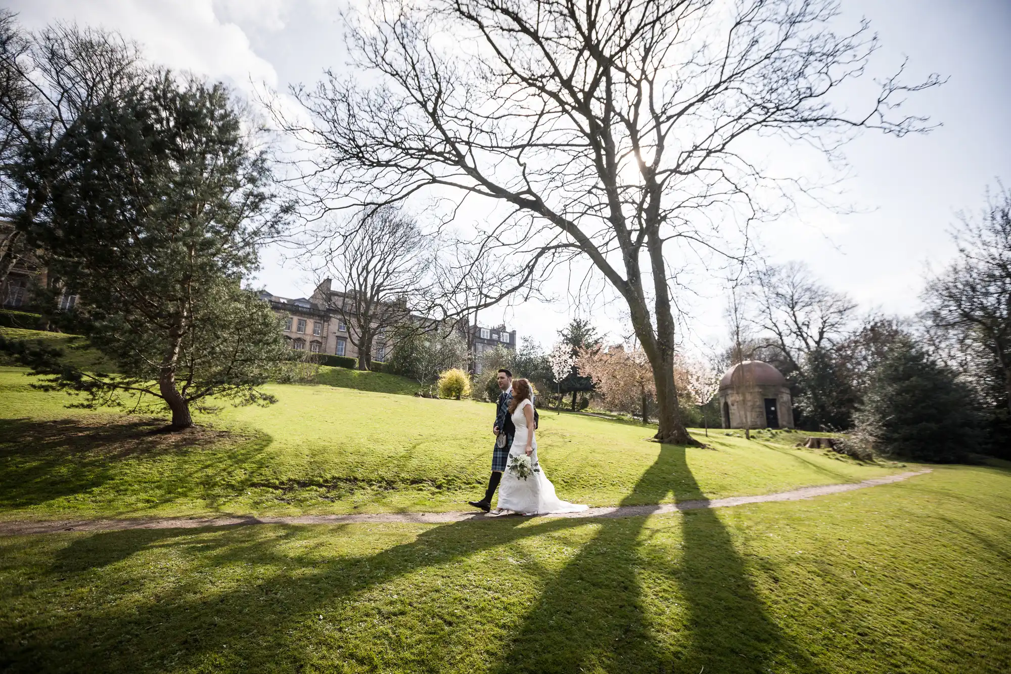 A bride and groom walk hand in hand on a narrow path through a sunlit park with trees and grass, shadows stretching across the ground, and buildings in the background.