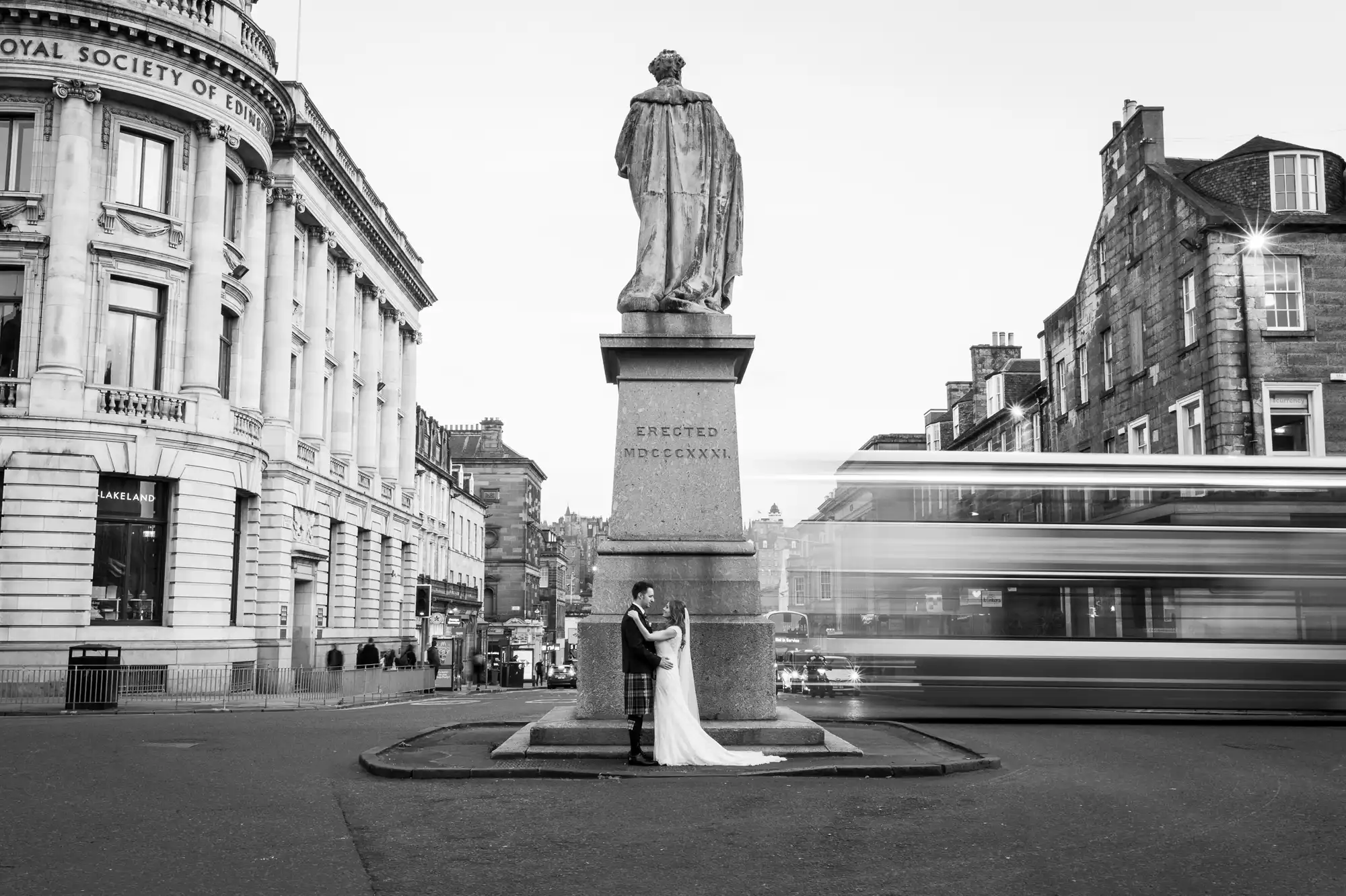 A bride and groom in wedding attire stand at the base of a large statue in a city street. A bus is captured in motion passing by them. Buildings surround the scene.