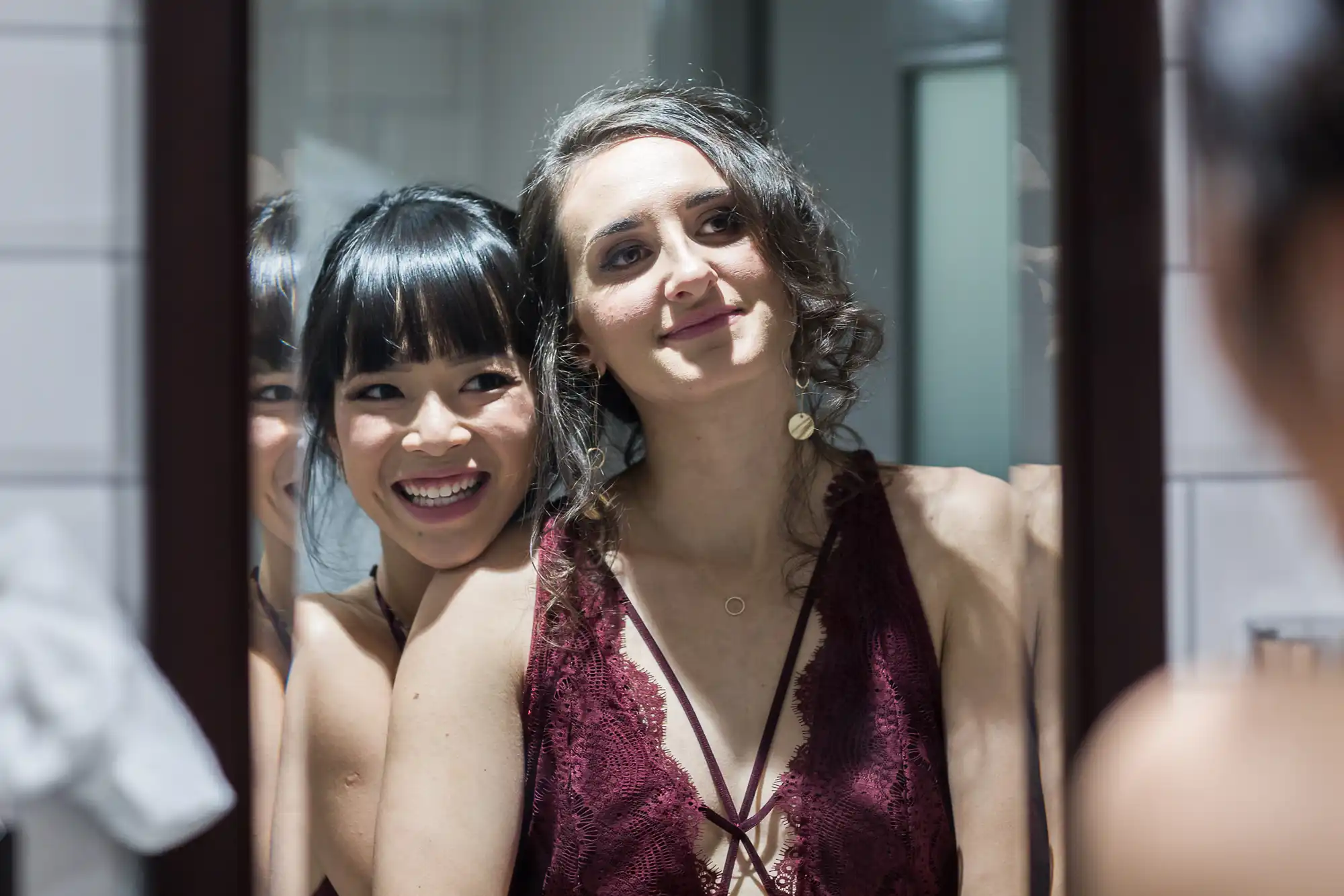Two women wearing maroon dresses smile as they look into a mirror in a well-lit room. One stands behind the other, embracing her from the side.