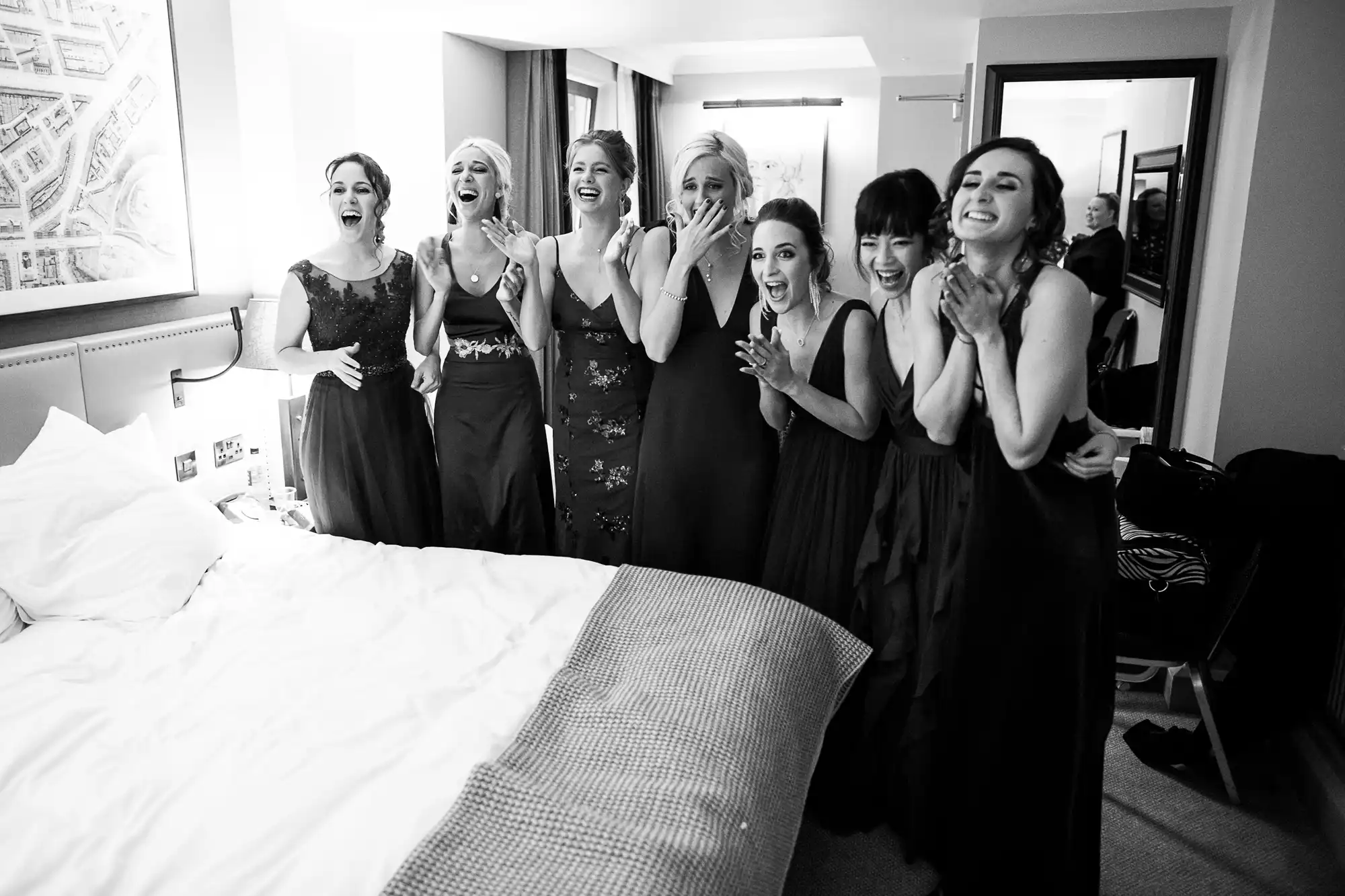 A group of seven bridesmaids in formal dresses stand close together in a bedroom, smiling and expressing excitement as they see the bride for the first time.