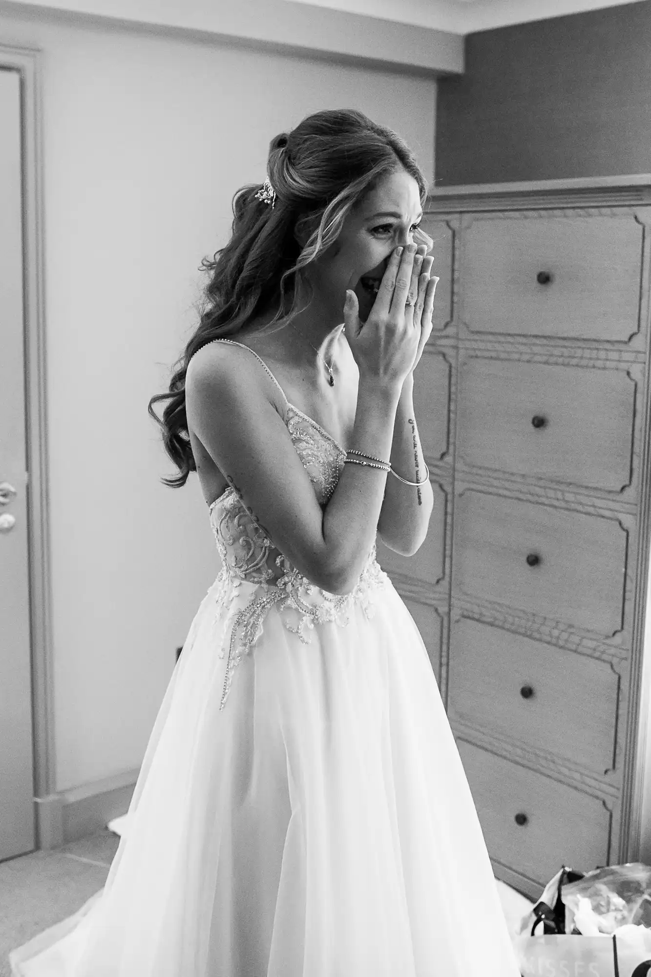 A woman in a white wedding dress stands indoors with hands covering her mouth, appearing emotional.