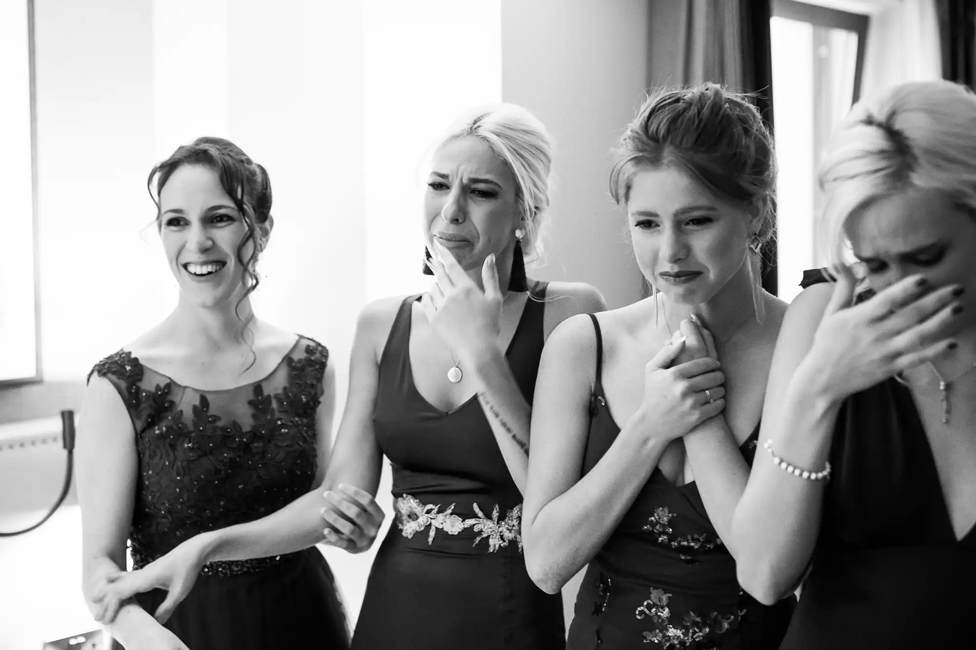 Four women stand close together; dressed in formal attire, three appear emotional, and one smiles. The scene is indoors, with natural light from a window.