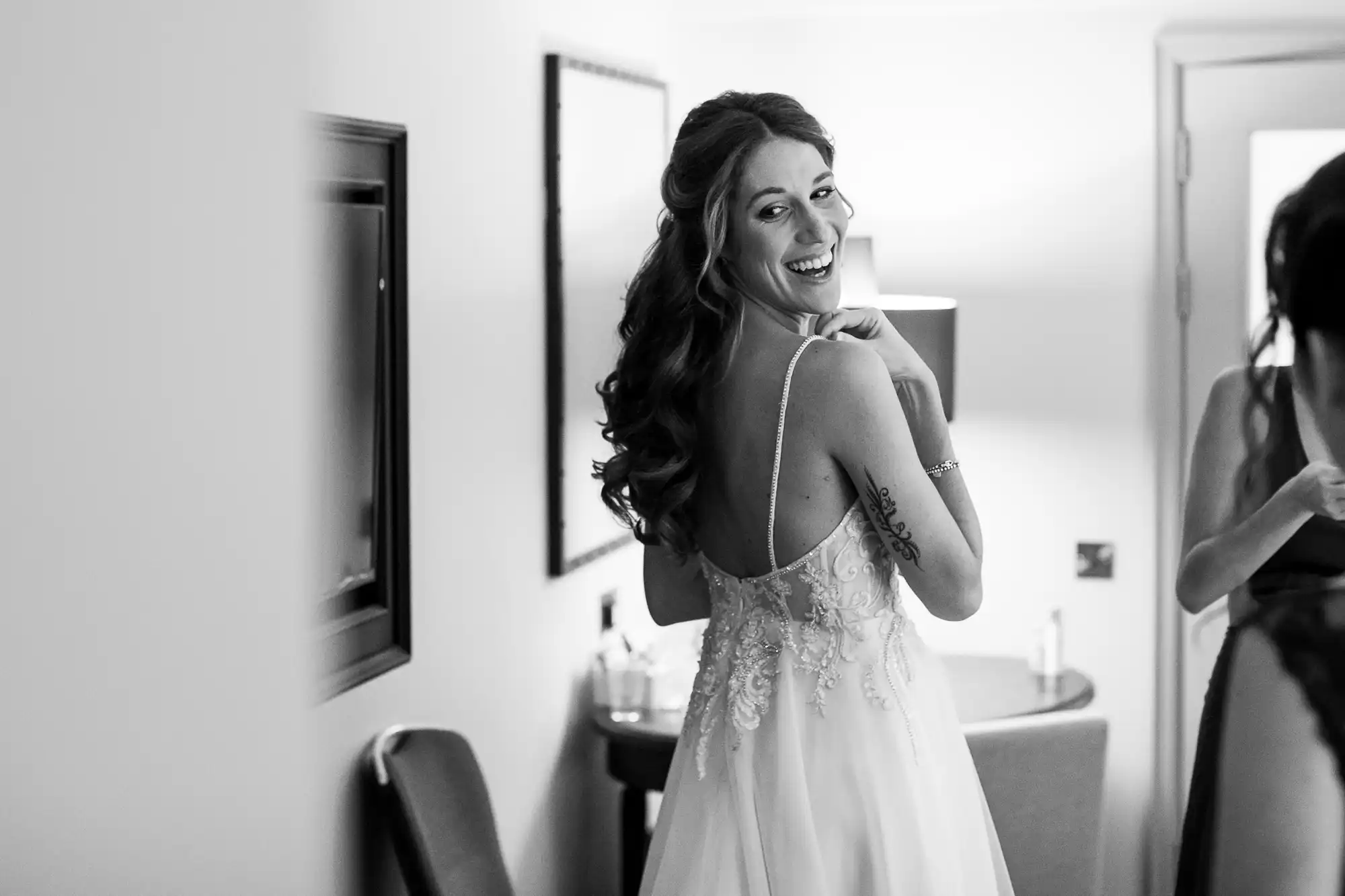A bride in a wedding dress looks over her shoulder and smiles in a well-lit room. Another person stands nearby, partly visible.