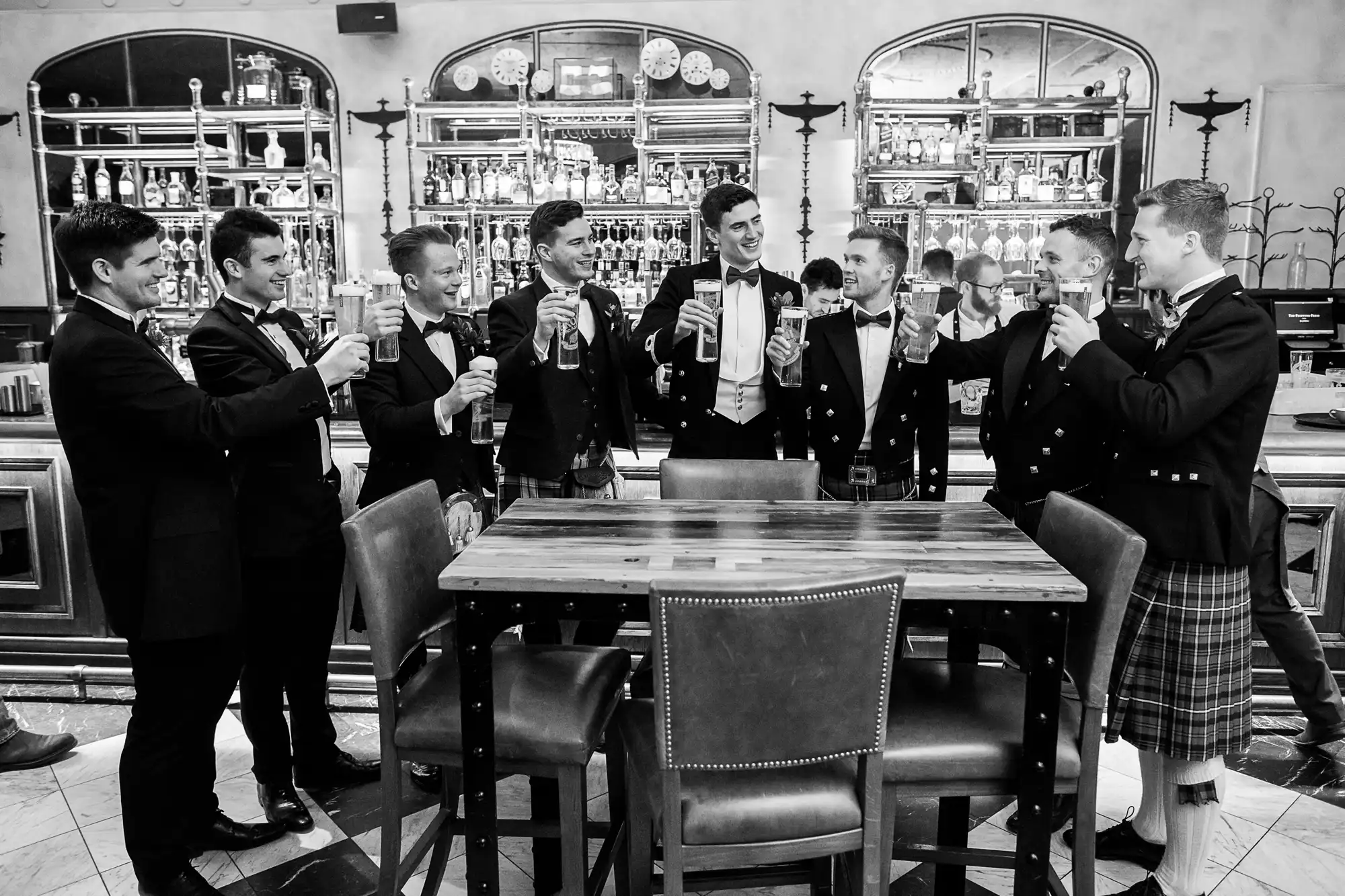 A group of men in formal attire, some wearing kilts, stand around a high table in a bar, raising their glasses in a celebratory toast.