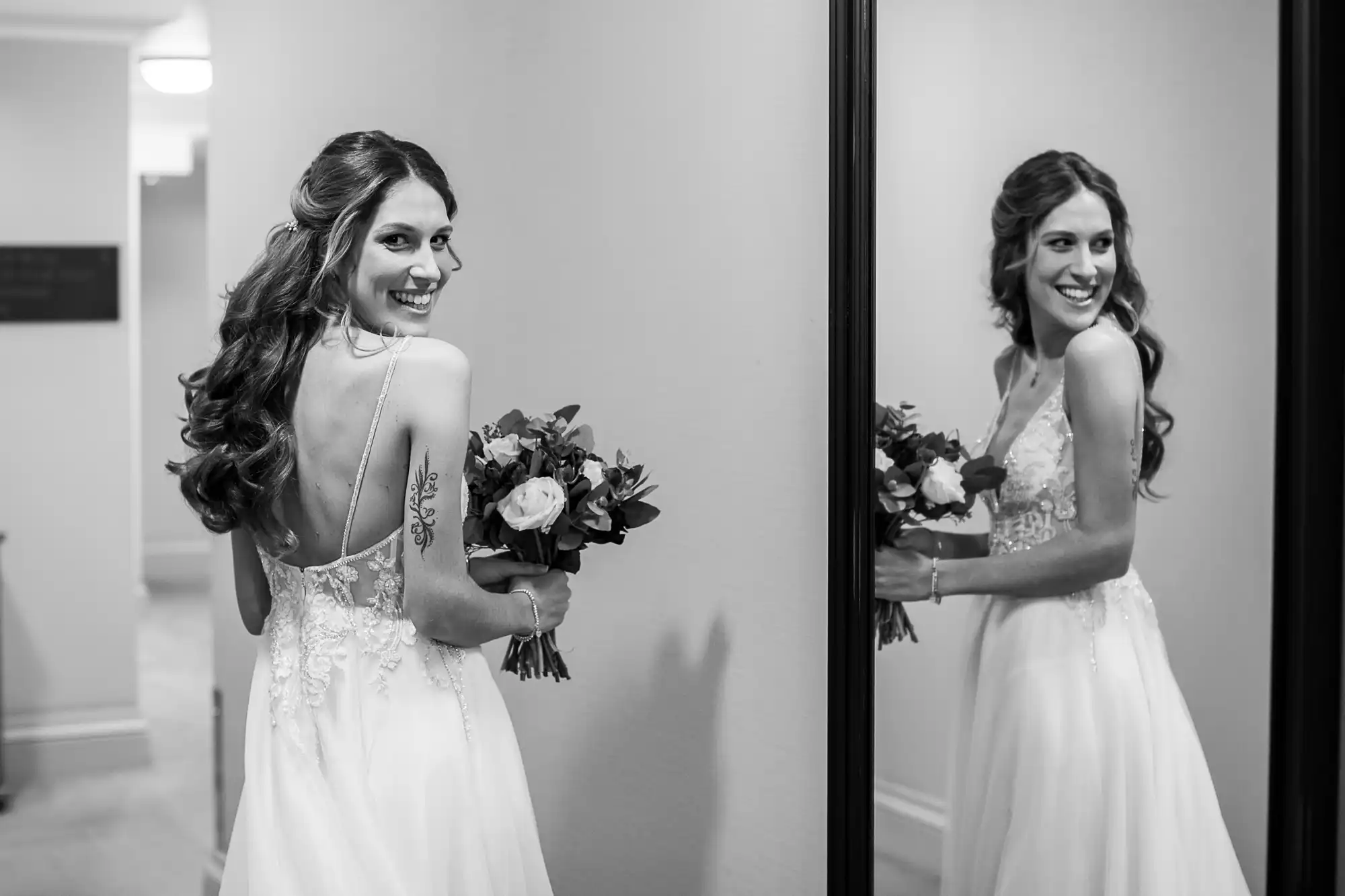 A bride in a sleeveless lace wedding dress holds a bouquet and smiles at her reflection in a large mirror.