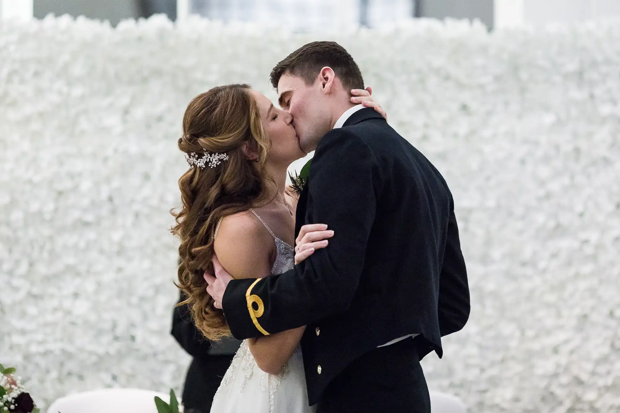 A bride and groom kiss passionately during their wedding ceremony. The bride wears a white dress and hair accessories; the groom is in a formal dark suit with gold trim. They embrace in front of a floral backdrop.