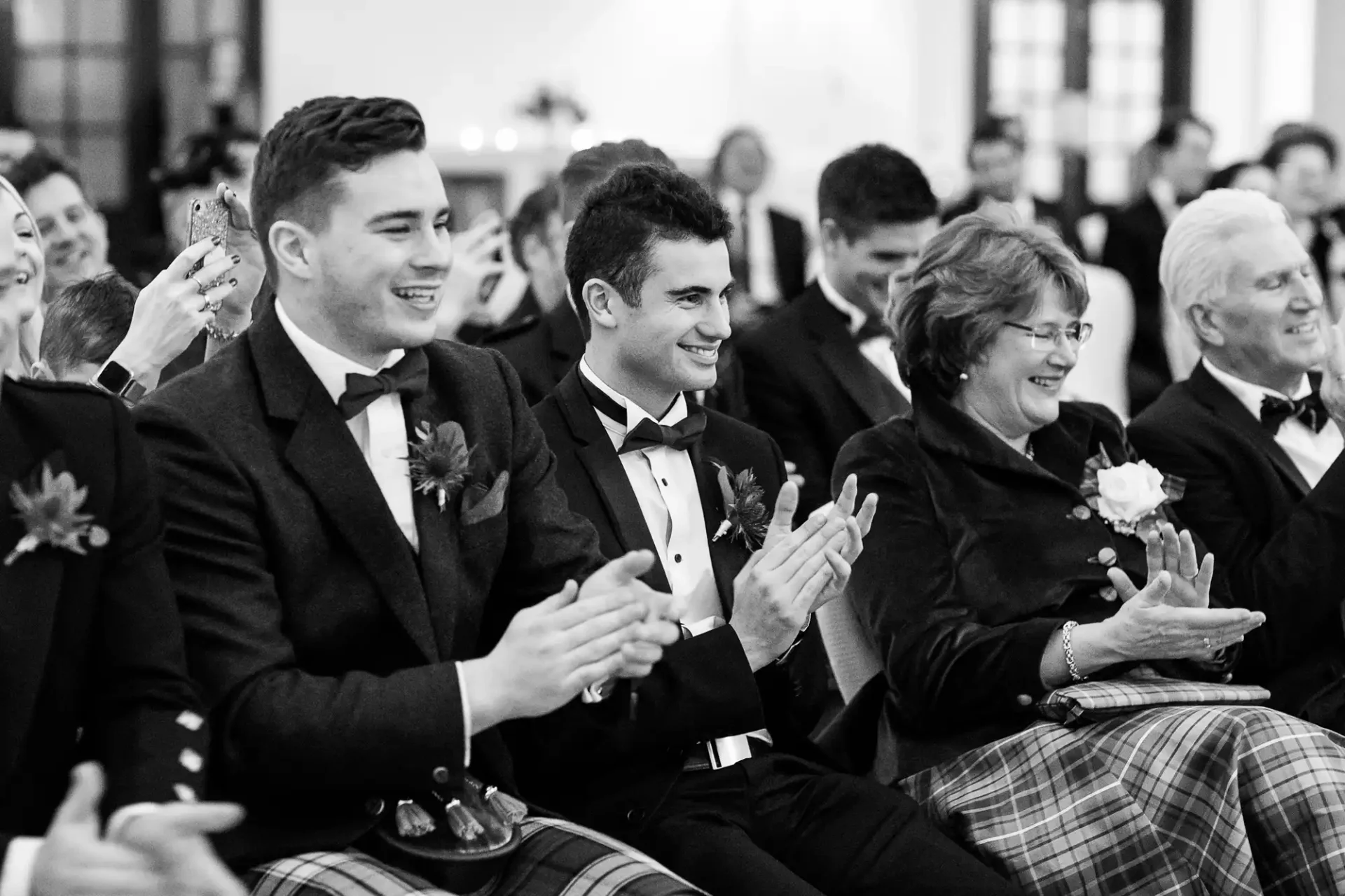 A group of people, dressed formally, sit and clap while smiling during an indoor event. The background is slightly out of focus.