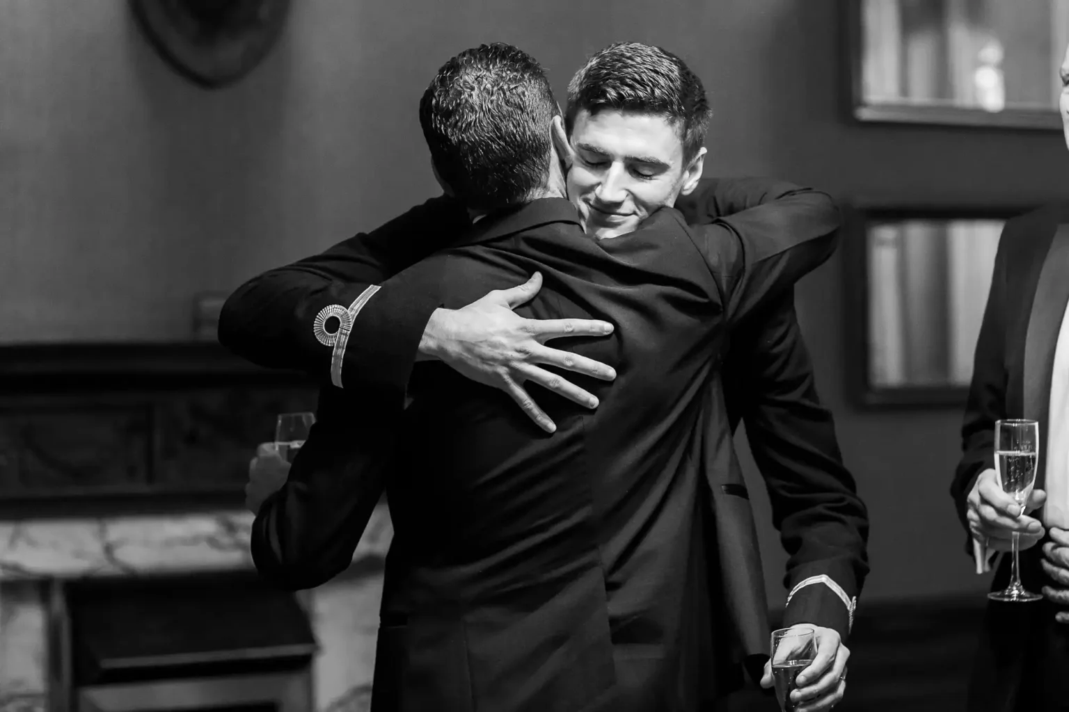 Two men in suits embrace warmly while holding drinks in a sophisticated setting. Both appear to be smiling, with one man seen from the back and the other facing the camera.