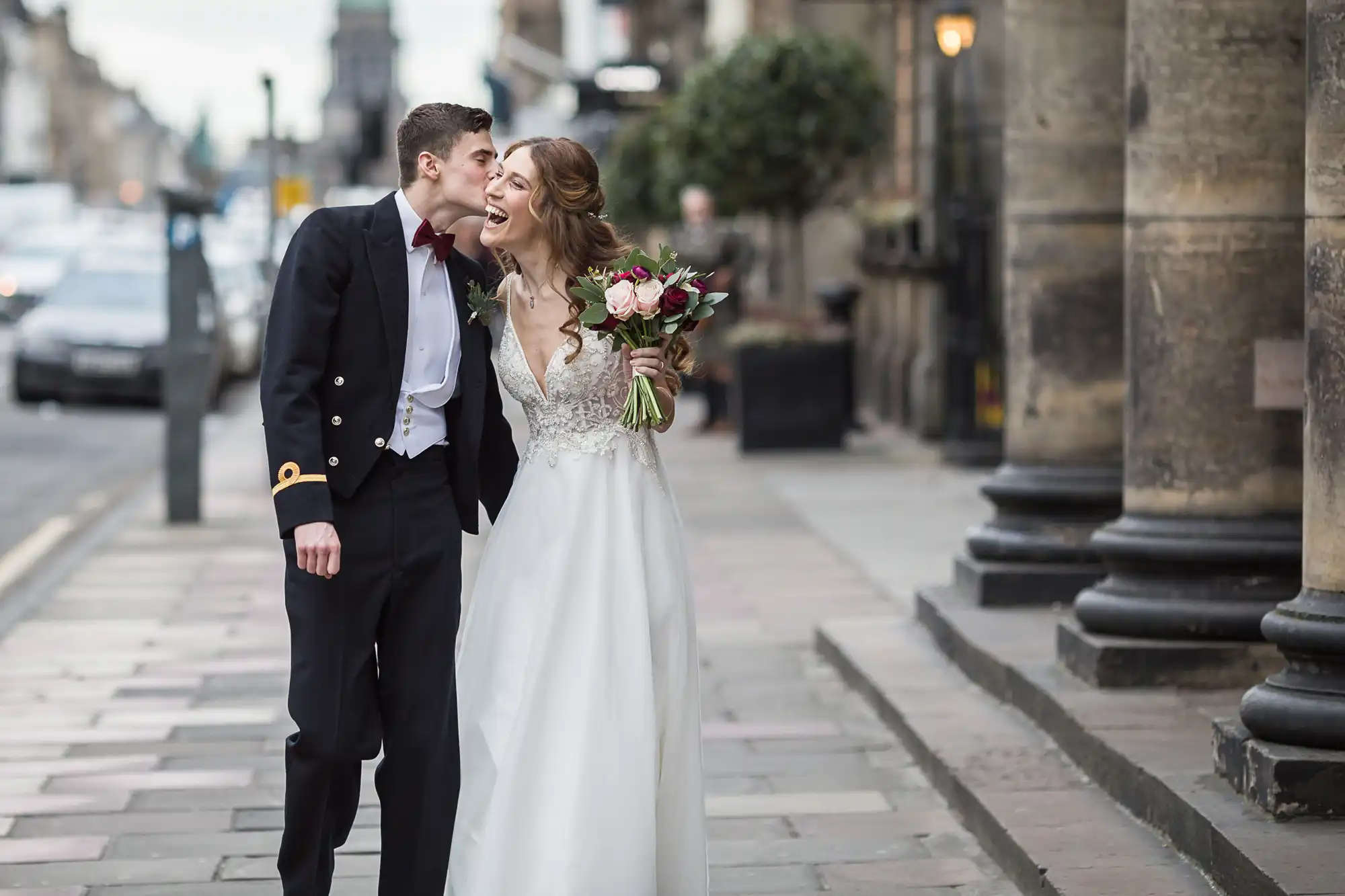 A bride and groom in formal attire walk on a city sidewalk; the groom kisses the bride on the forehead while she holds a bouquet and smiles.