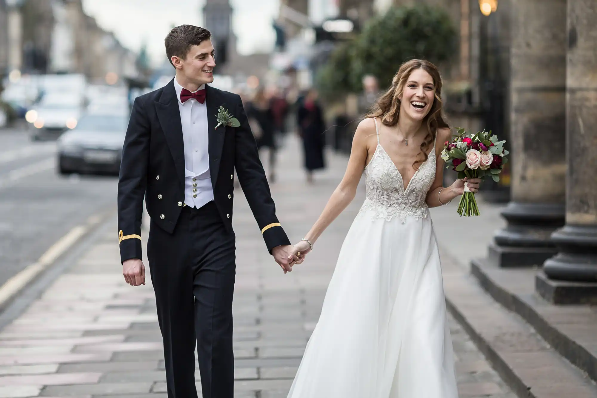 A bride in a white gown and a groom in a black tuxedo walk hand in hand on a city sidewalk, both smiling.