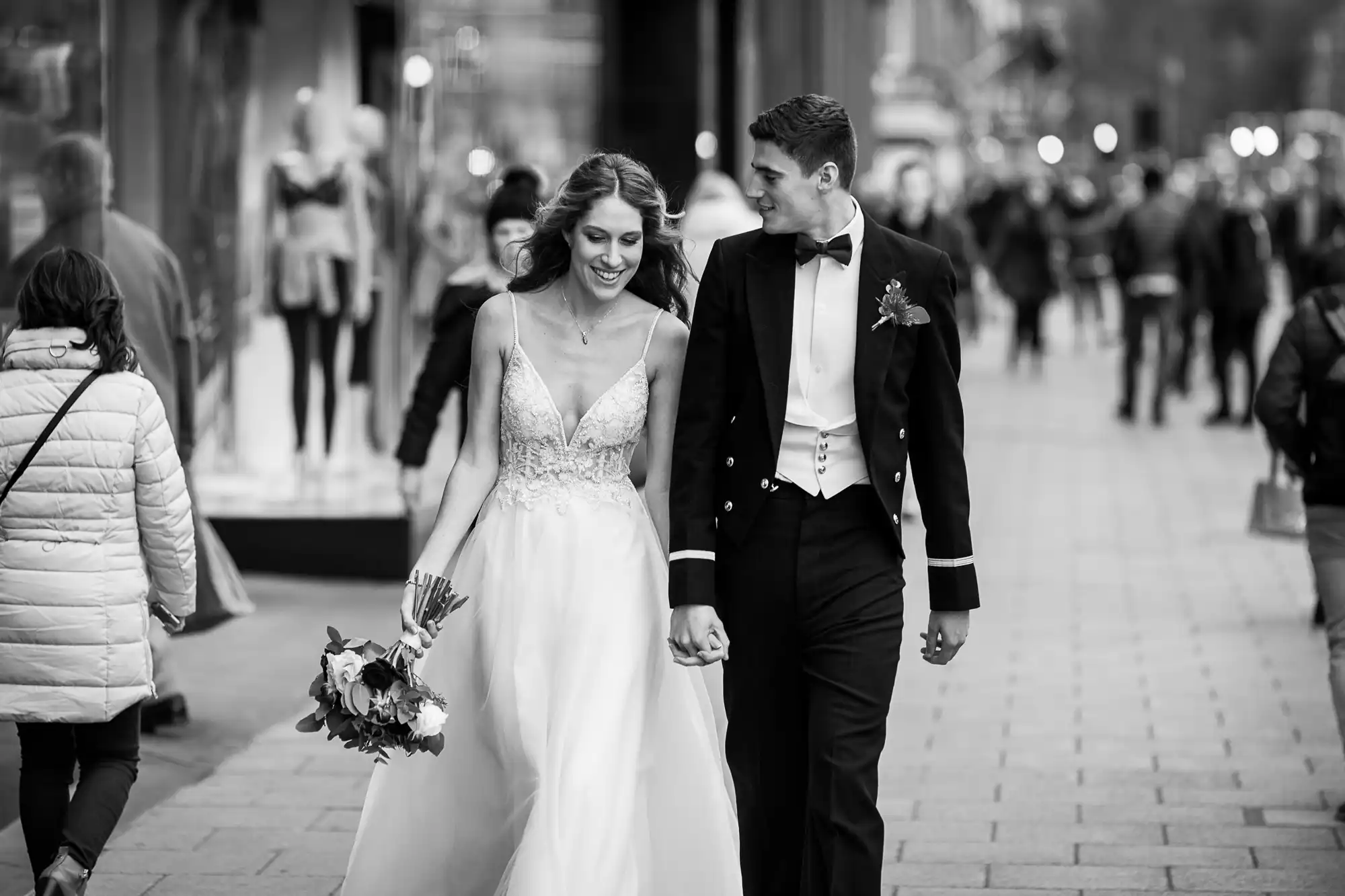 A bride and groom walk hand-in-hand down a busy city street, smiling at each other. The bride holds a bouquet, wearing a wedding dress, while the groom wears a suit and boutonniere.