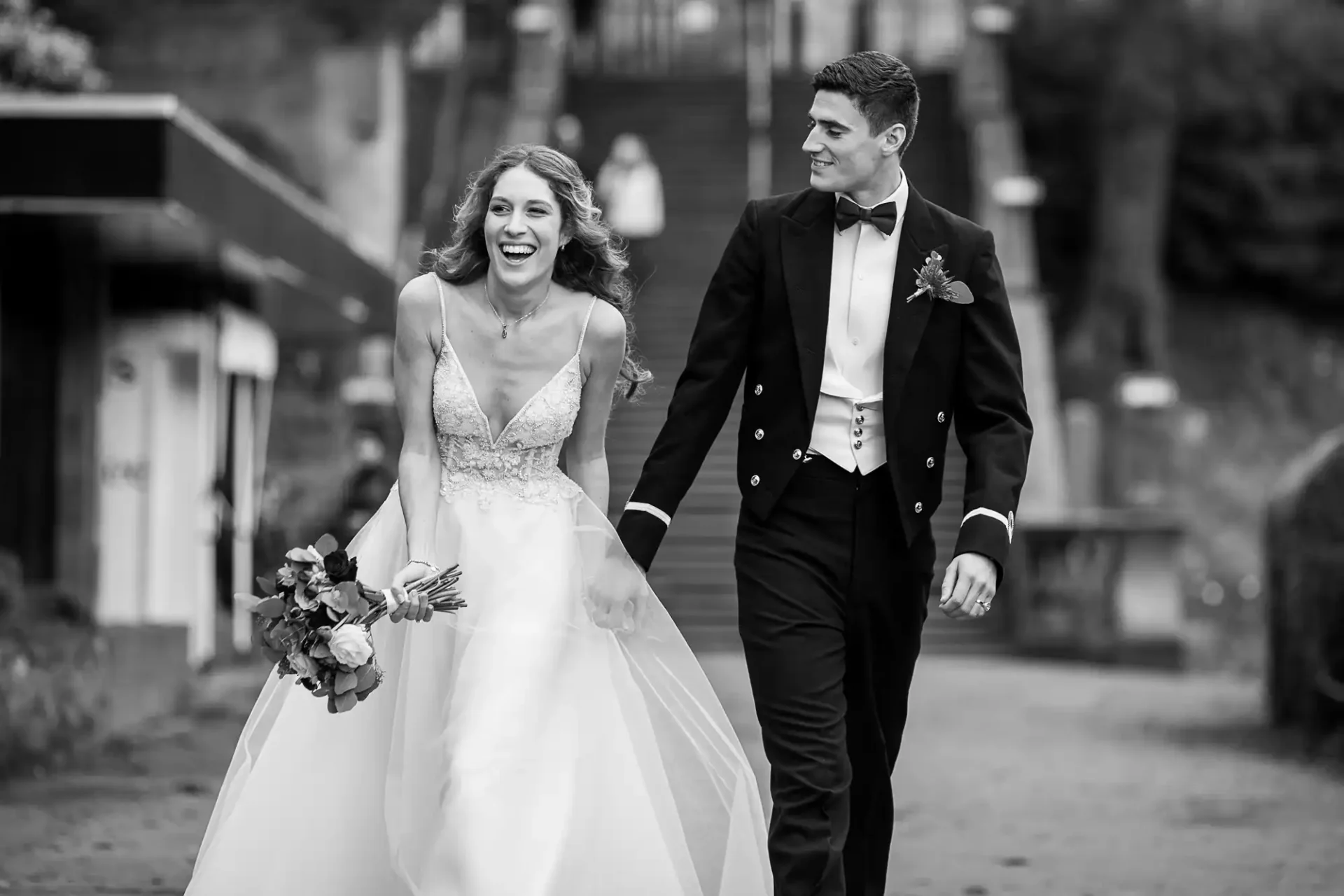 A bride and groom dressed in wedding attire hold hands and walk outside, both smiling and looking at each other. The bride holds a bouquet of flowers. Stairs and trees are visible in the background.