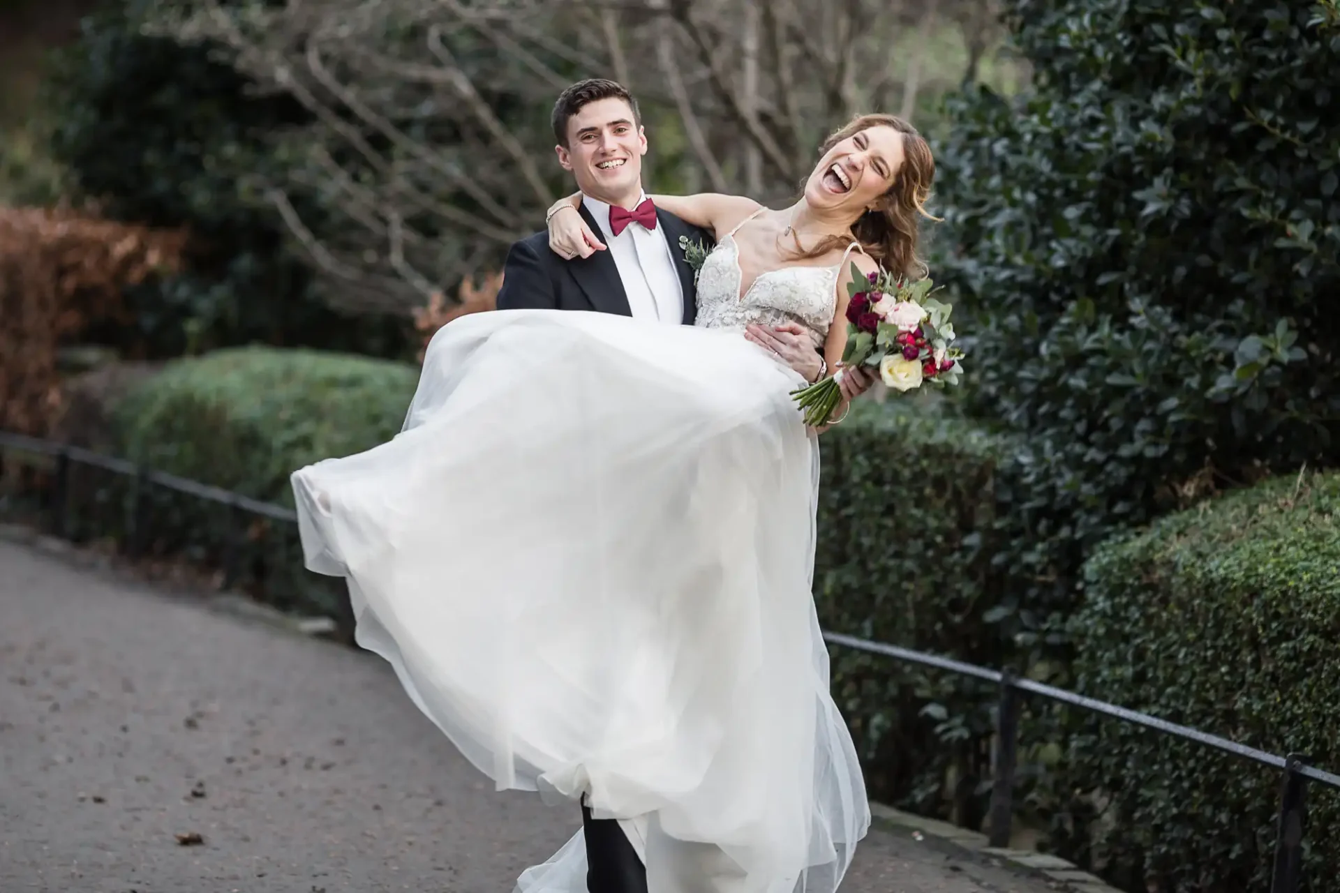 A groom in a black suit and red bow tie carries a smiling bride in a white wedding dress as they walk outdoors, surrounded by greenery.