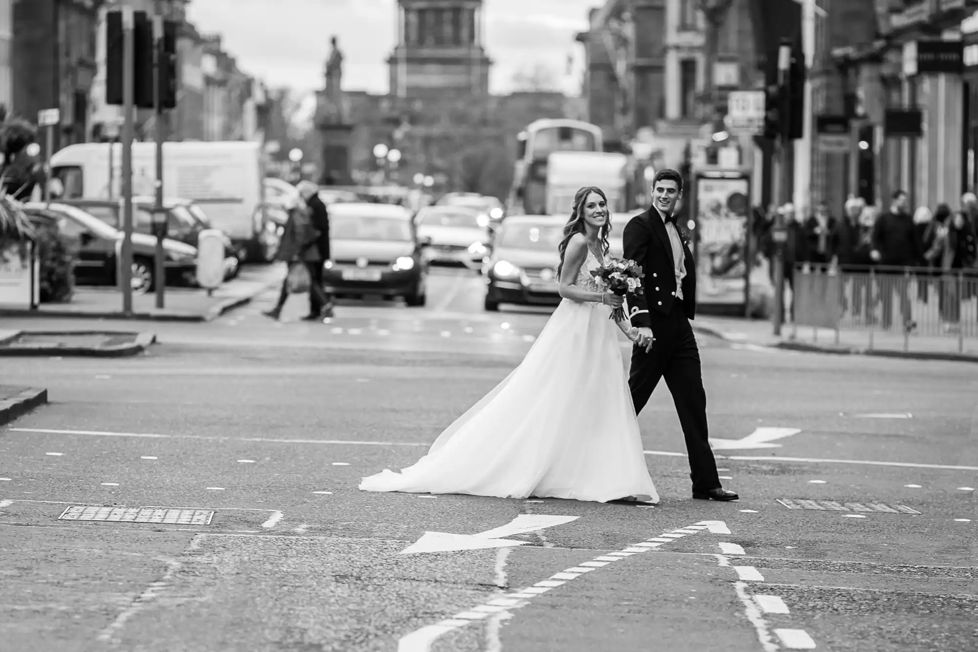 A bride and groom walk hand in hand across a busy street in a city, with cars, buses, and pedestrians in the background.