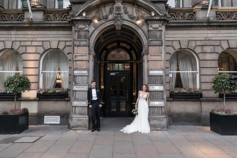 George Hotel newlyweds Paula and Alex: A couple, dressed in formal wedding attire, stands in front of an ornate building entrance. The groom is on the left, leaning against a pillar, and the bride is on the right, holding a bouquet.