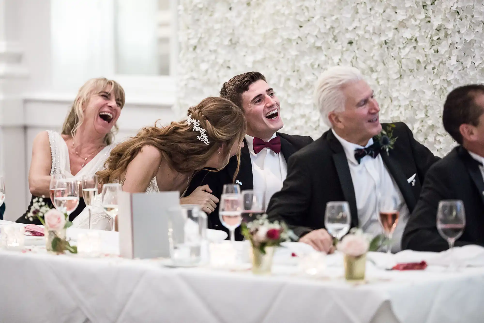 A group of people dressed in formal attire are seated at a table, laughing and looking towards the left. The background features a white floral wall.