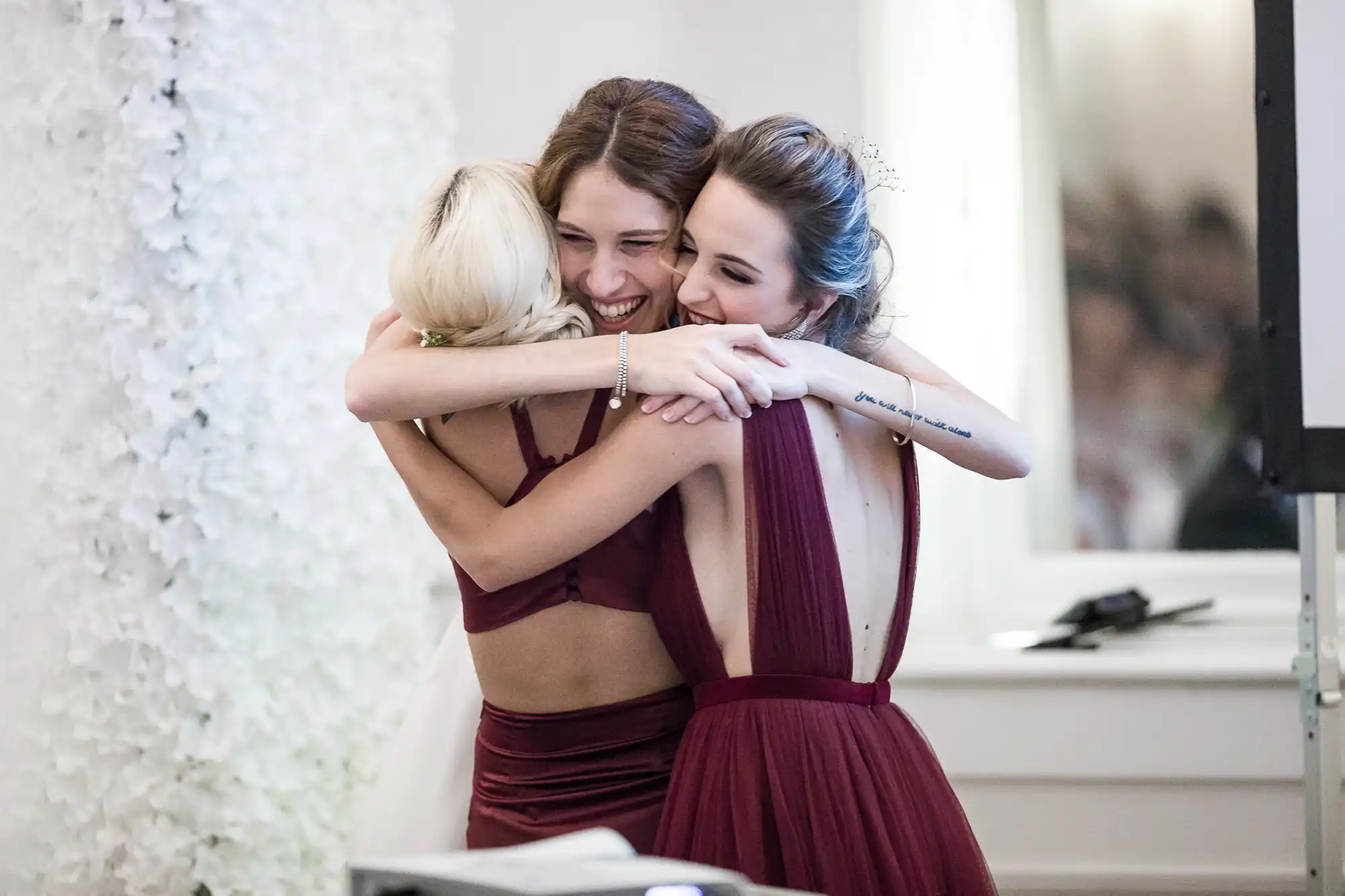 Bride and bridesmaids in formal dresses share a group hug, smiling and celebrating indoors with a white floral backdrop.
