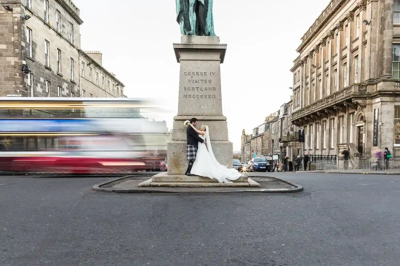 Edinburgh George Hotel wedding: A bride and groom stand in front of a statue on a street island, while a red and white bus passes by in the background.