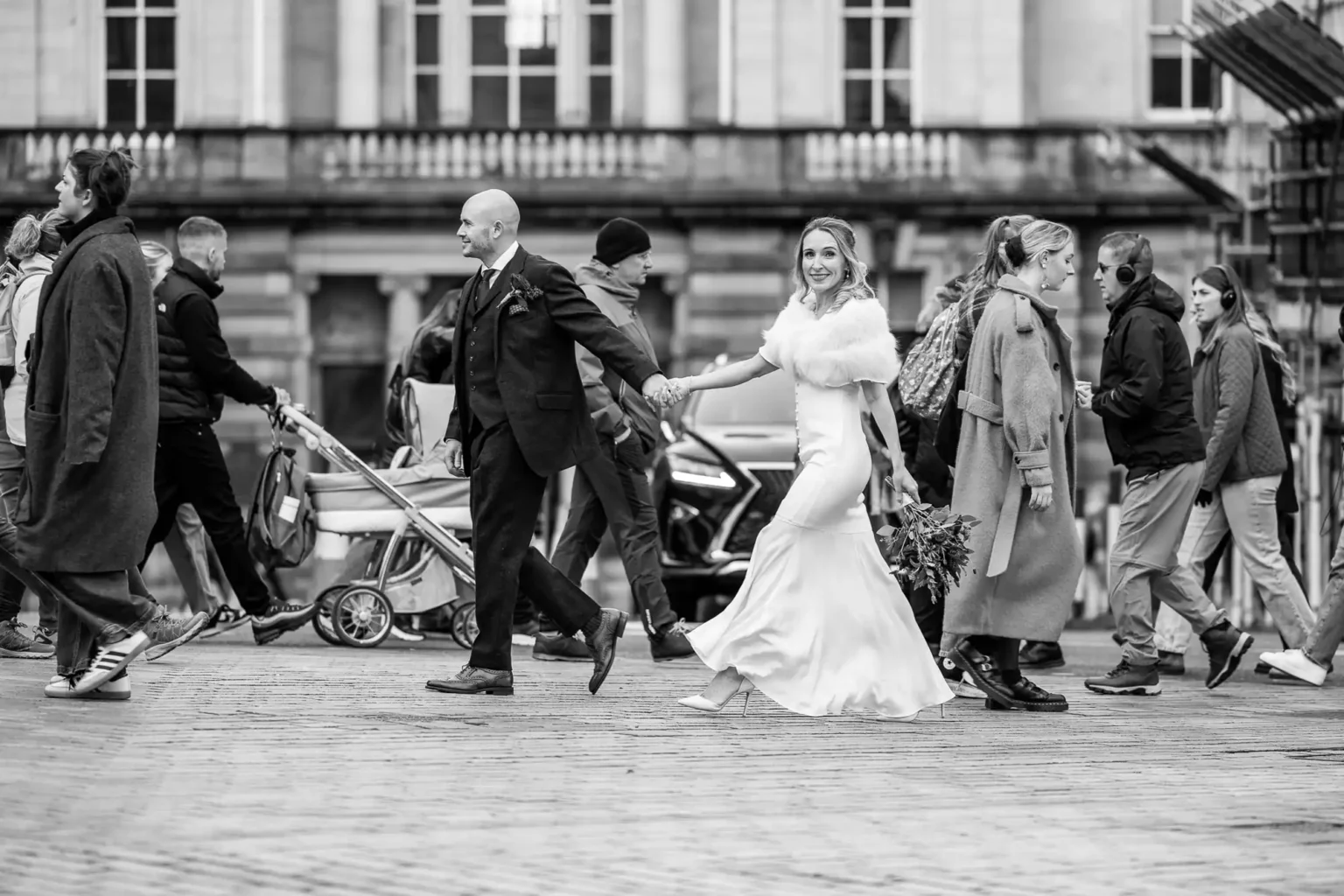 A bride and groom walk hand in hand across a busy city street while pedestrians pass by in both directions.