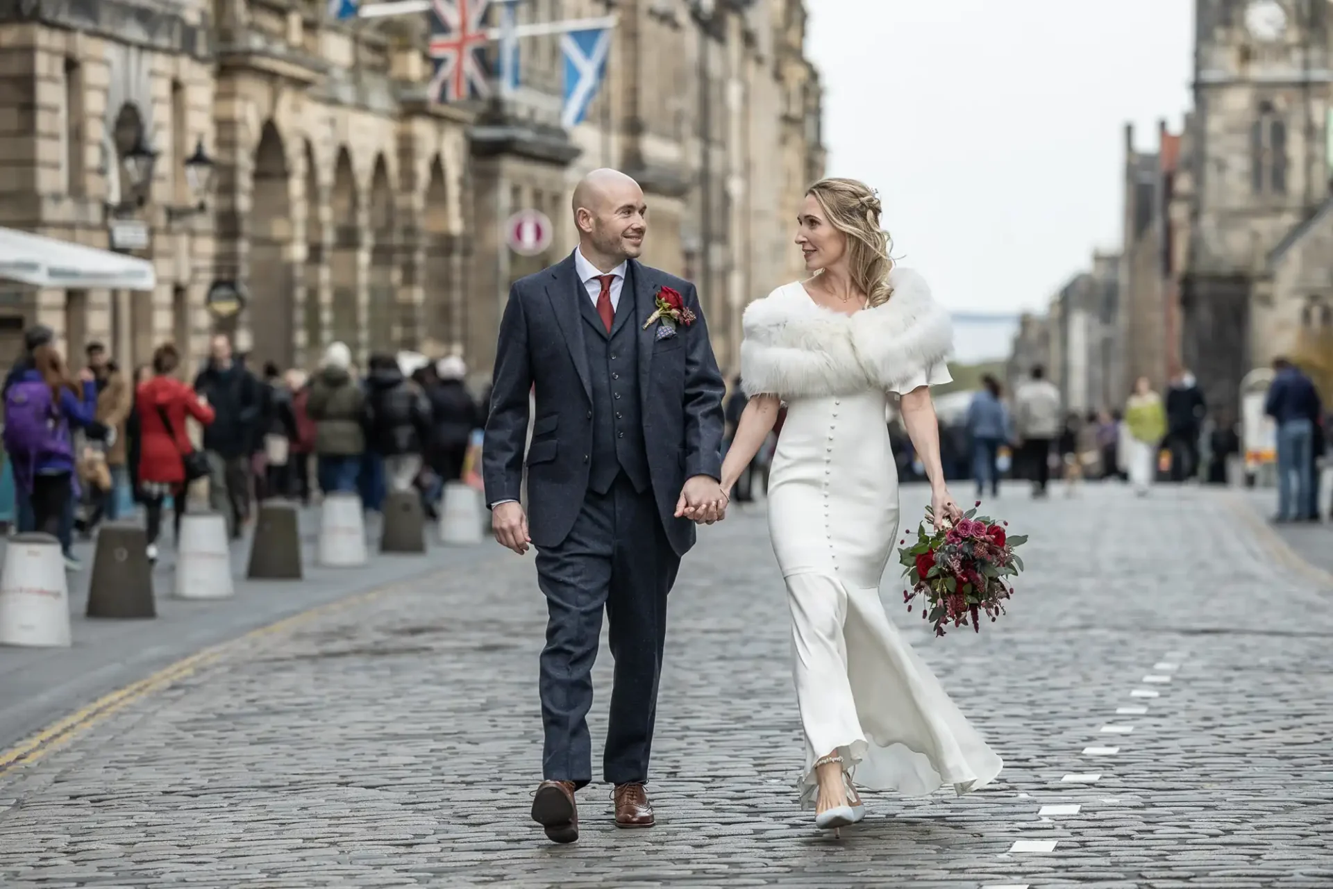 A bride and groom walk hand in hand down a cobblestone street, dressed in formal wedding attire, with onlookers in the background.
