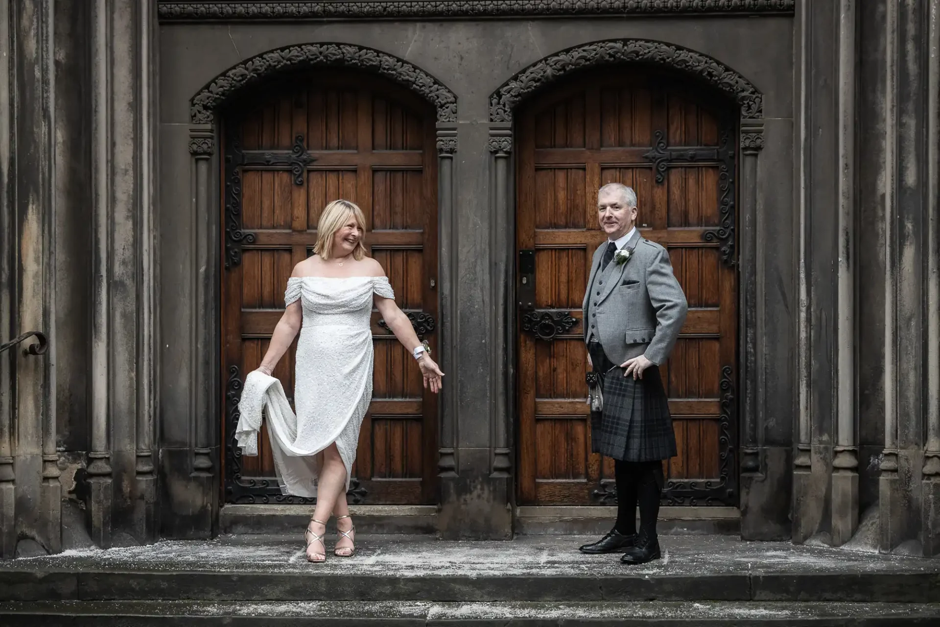 A woman in a white dress and a man in a kilt stand on stone steps in front of two large wooden doors, posing for a photo.