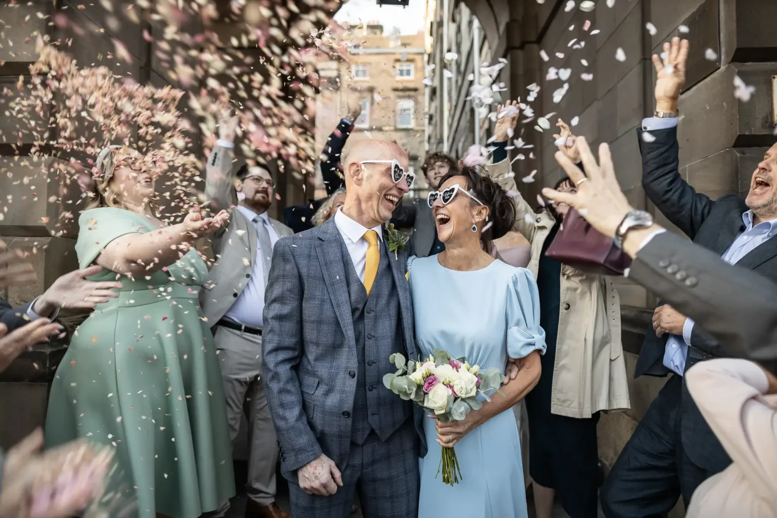 A newlywed couple smiles and walks through a crowd of people throwing confetti. The groom wears a gray suit and the bride holds a bouquet and wears a light blue dress.