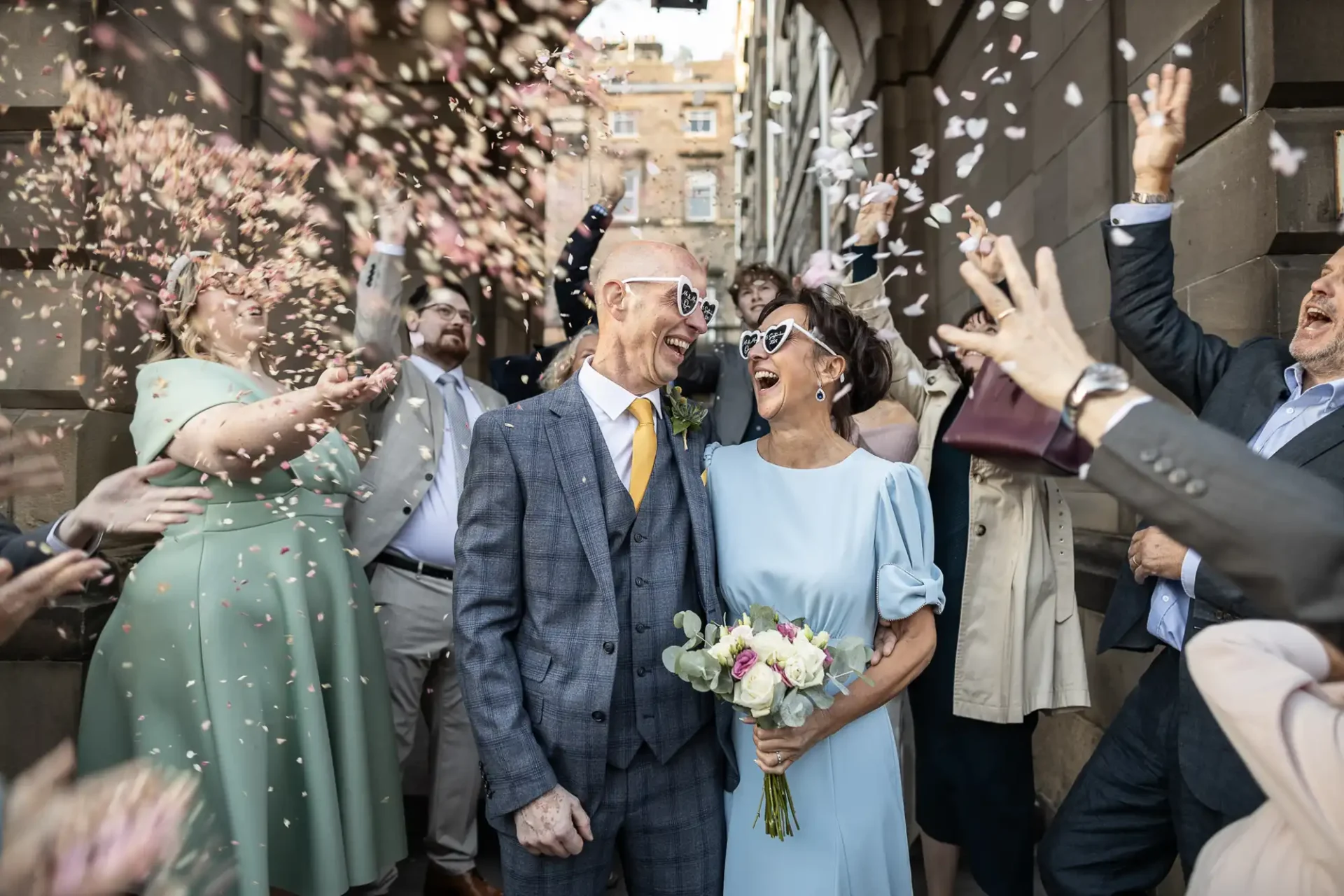 A newlywed couple smiles and walks through a crowd of people throwing confetti. The groom wears a gray suit and the bride holds a bouquet and wears a light blue dress.