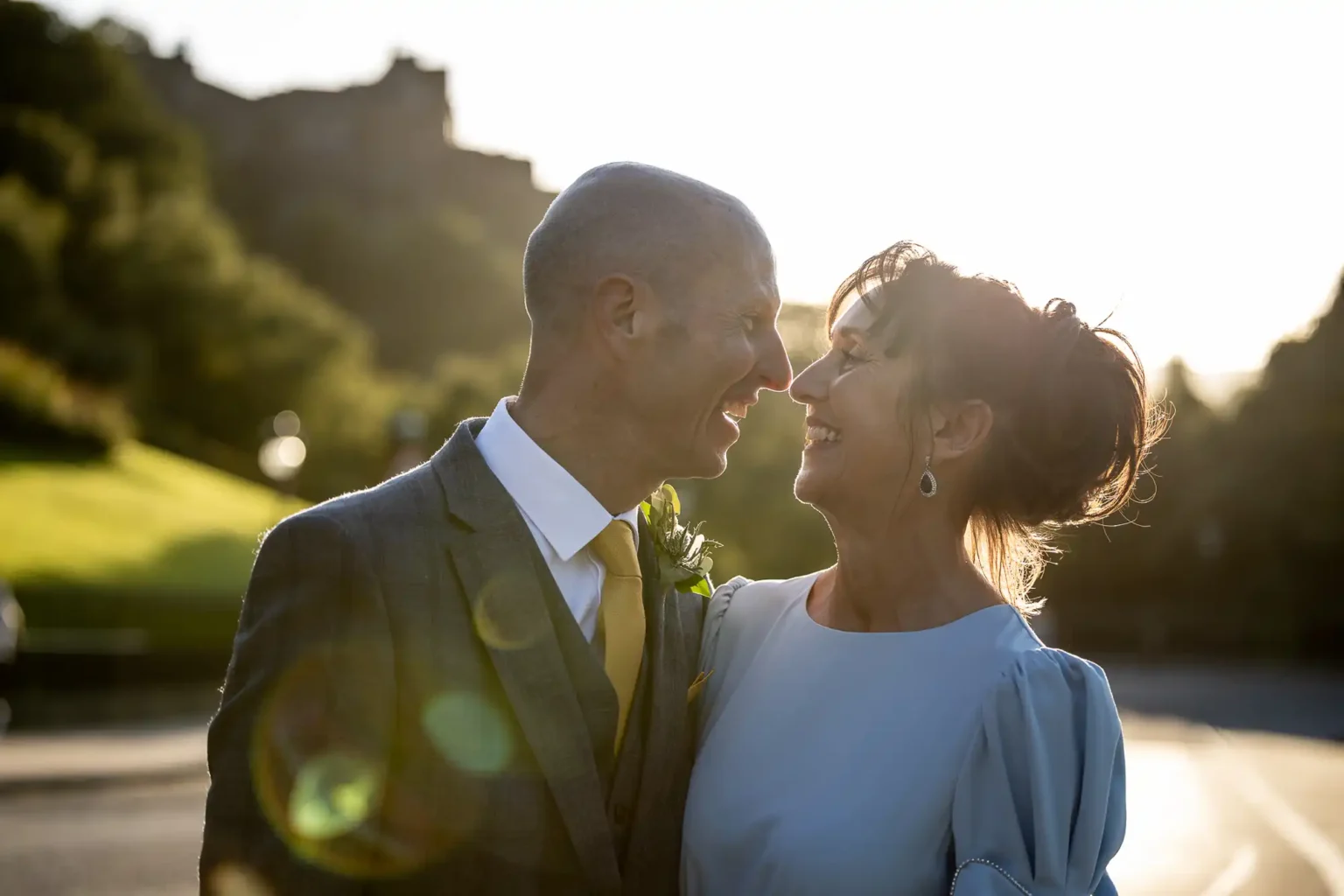 A couple dressed formally stands outside, smiling at each other in the sunlight with greenery and a blurred building in the background.