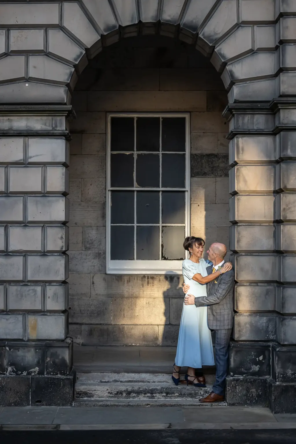 An older couple stands embracing under a stone archway, illuminated by sunlight, with a large window behind them on the building’s wall.