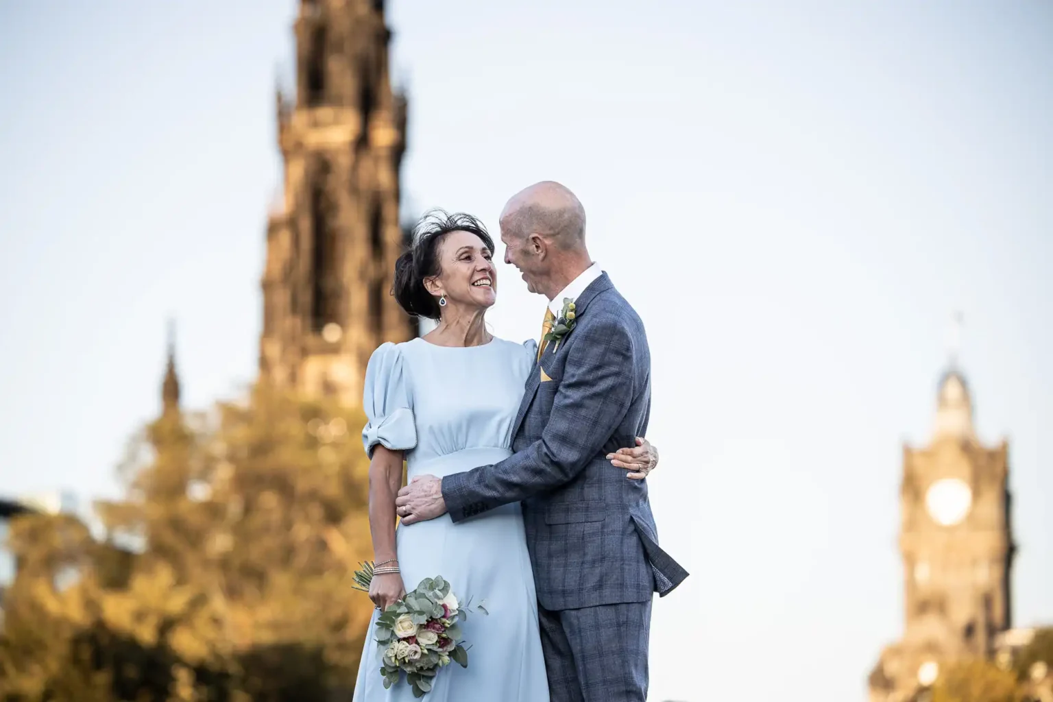 An older couple dressed formally, with the woman holding a bouquet, stand close together outdoors in front of historic towers, smiling at each other.