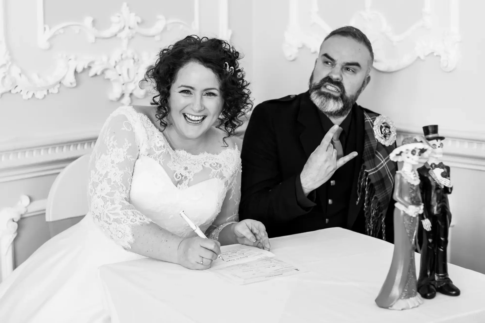 A bride smiles while signing a document at a table. Next to her, the groom makes a hand gesture and wears a suit with a tartan sash. A wedding figurine is also on the table.