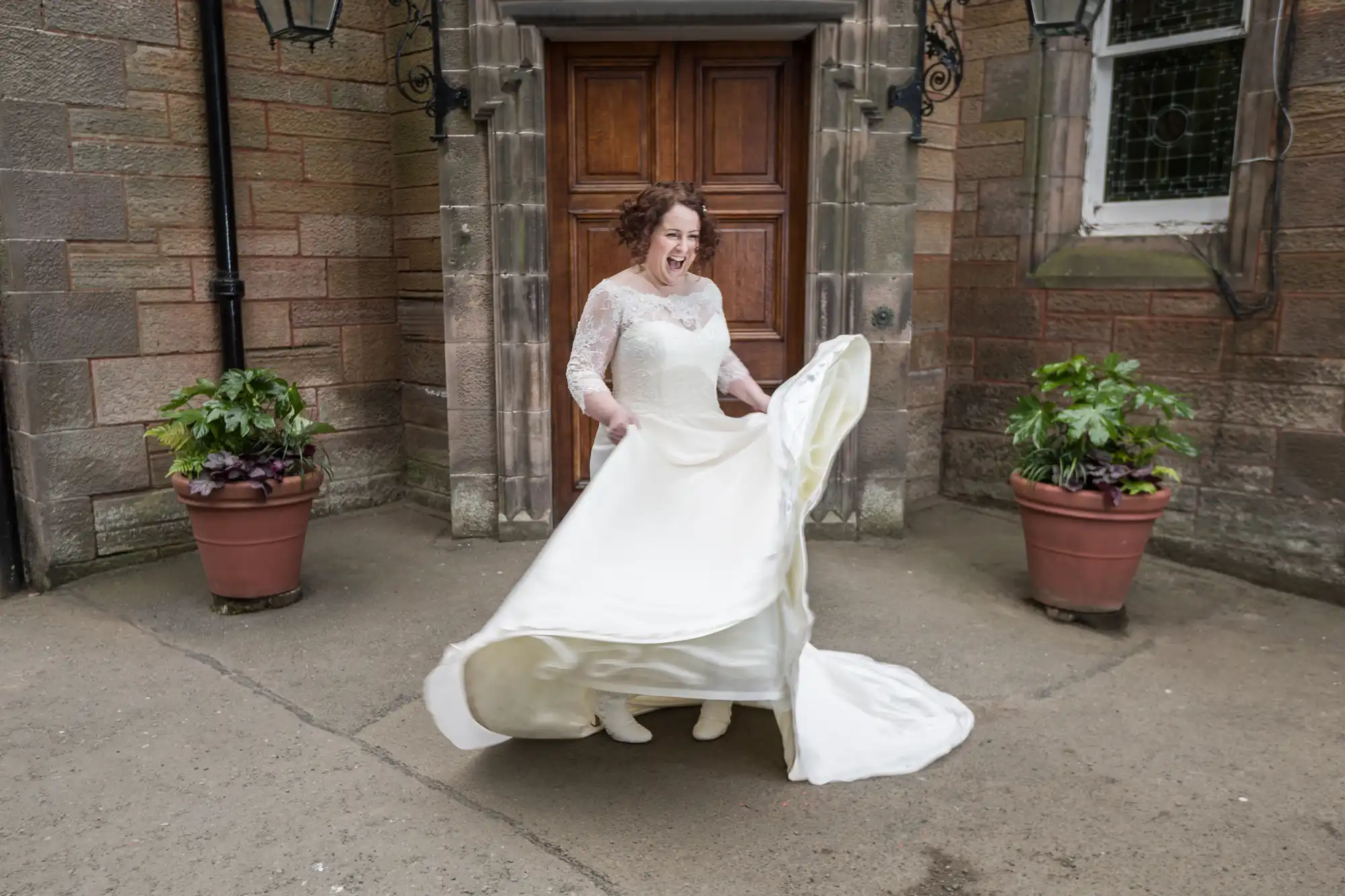A bride wearing a wedding dress twirls joyfully in front of a wooden door, flanked by potted plants on a stone patio.