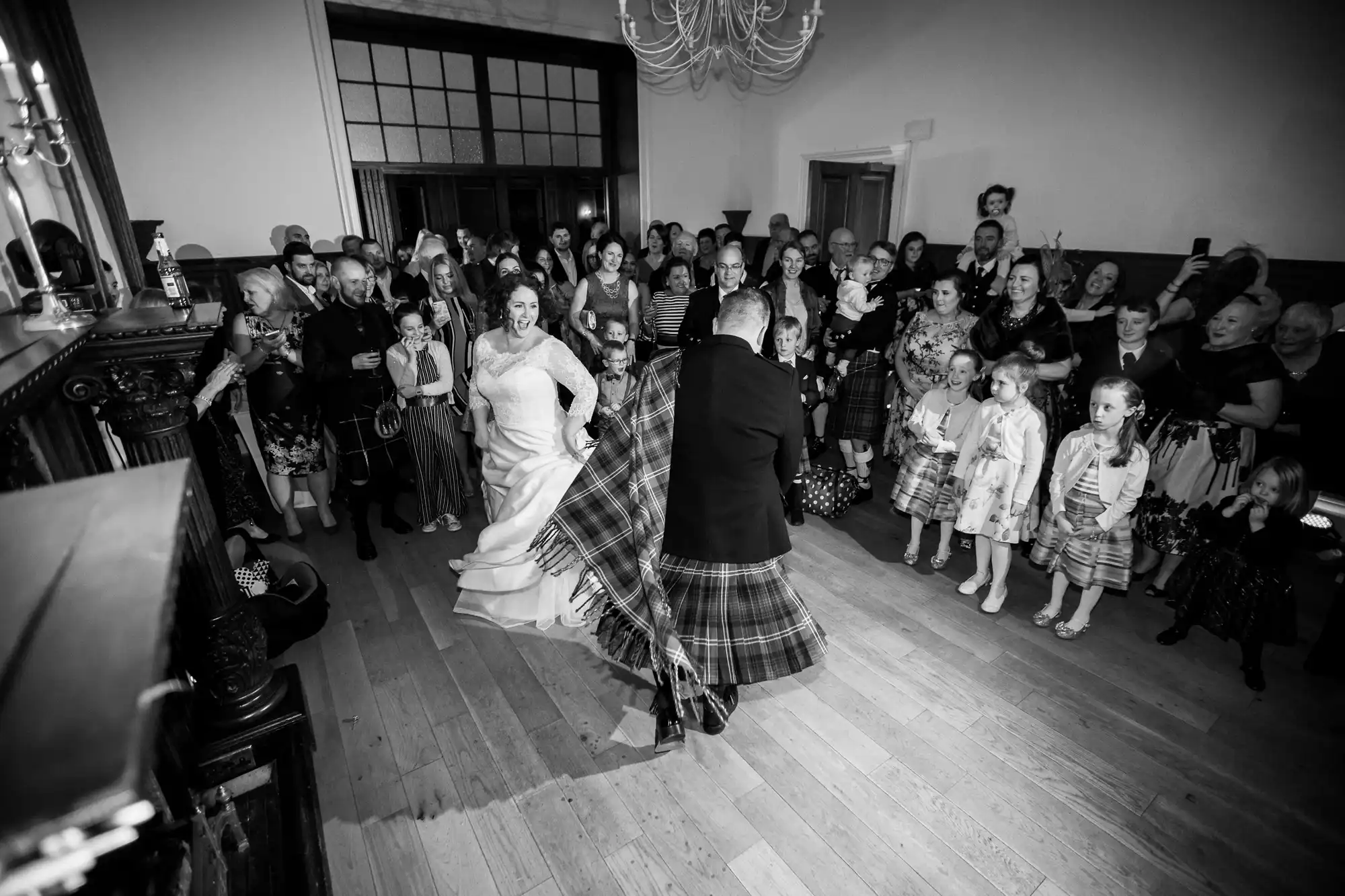 A bride and groom dance in a room filled with guests. Many attendees, including children, wear traditional Scottish attire. The setting is a wooden floored hall with large windows and a chandelier.