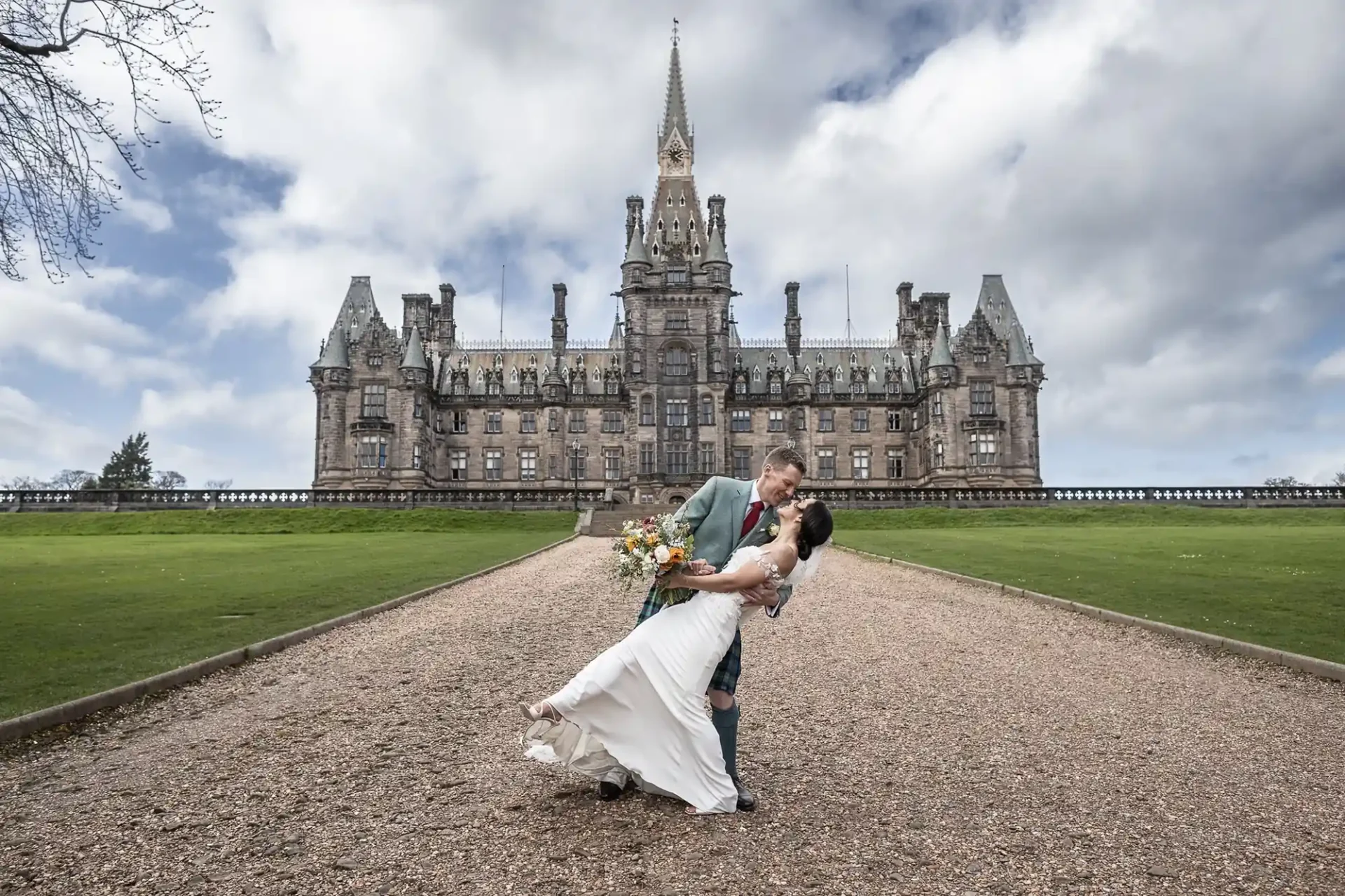 A newlywed couple poses on a gravel path in front of Fettes College under a cloudy sky.