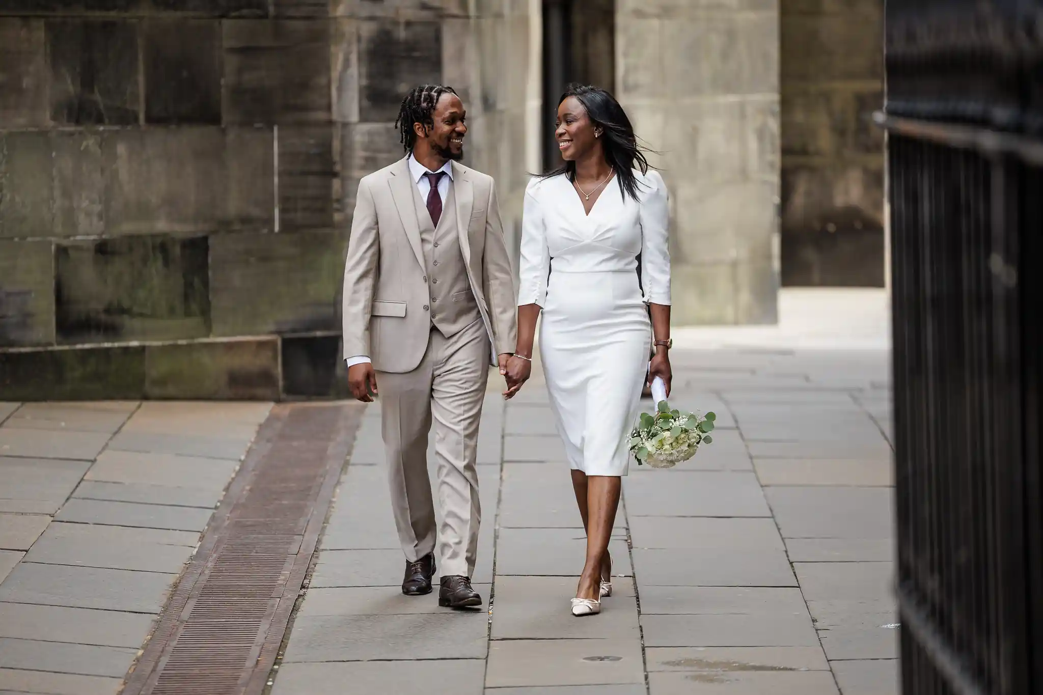 A couple dressed in formal attire, holding hands and walking outdoors on a stone pathway. The woman holds a bouquet of flowers.
