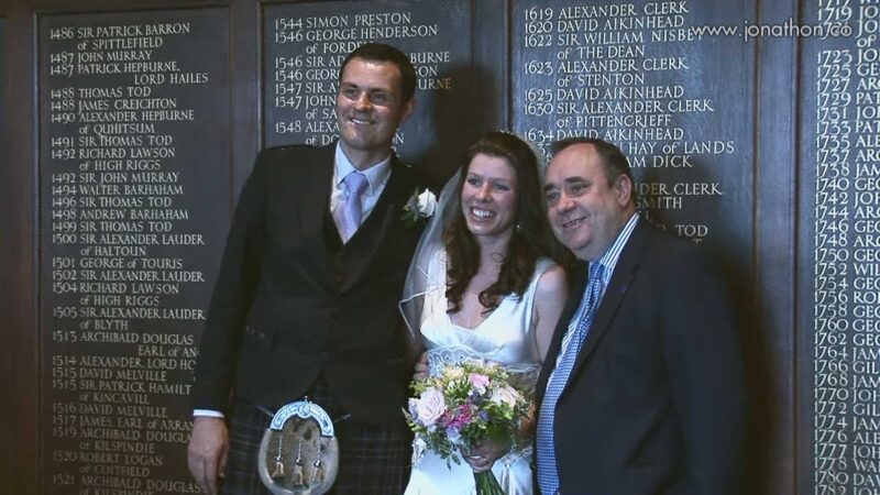 Three people pose together in front of a dark wooden wall with engraved names. Two men and one woman stand smiling, with the woman holding a bouquet of flowers - First Minister Alex Salmond meets newlyweds.